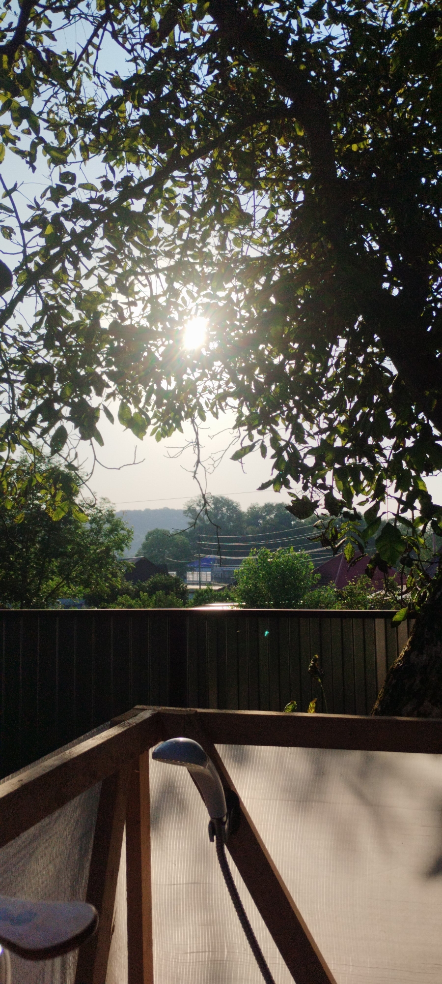 The image depicts a sunlit outdoor scene, likely a backyard or garden area, seen through the branches of a tree. In the foreground, a wooden structure with a translucent plastic sheeting forms a sort of enclosure or shelter. A shower head is mounted within this structure, attached to a flexible hose. Beyond this, a dark-colored corrugated metal fence runs horizontally across the frame, separating the foreground from the background. The background reveals a hazy landscape with green trees, some buildings, and power lines, suggesting a semi-rural or suburban setting. The sun is shining brightly, creating lens flare and casting shadows. The overall impression is one of a tranquil, possibly rustic, outdoor space. The location is specified as Yaroslavskaya, Russia, implying a setting within that region. There are no people visible in the image.