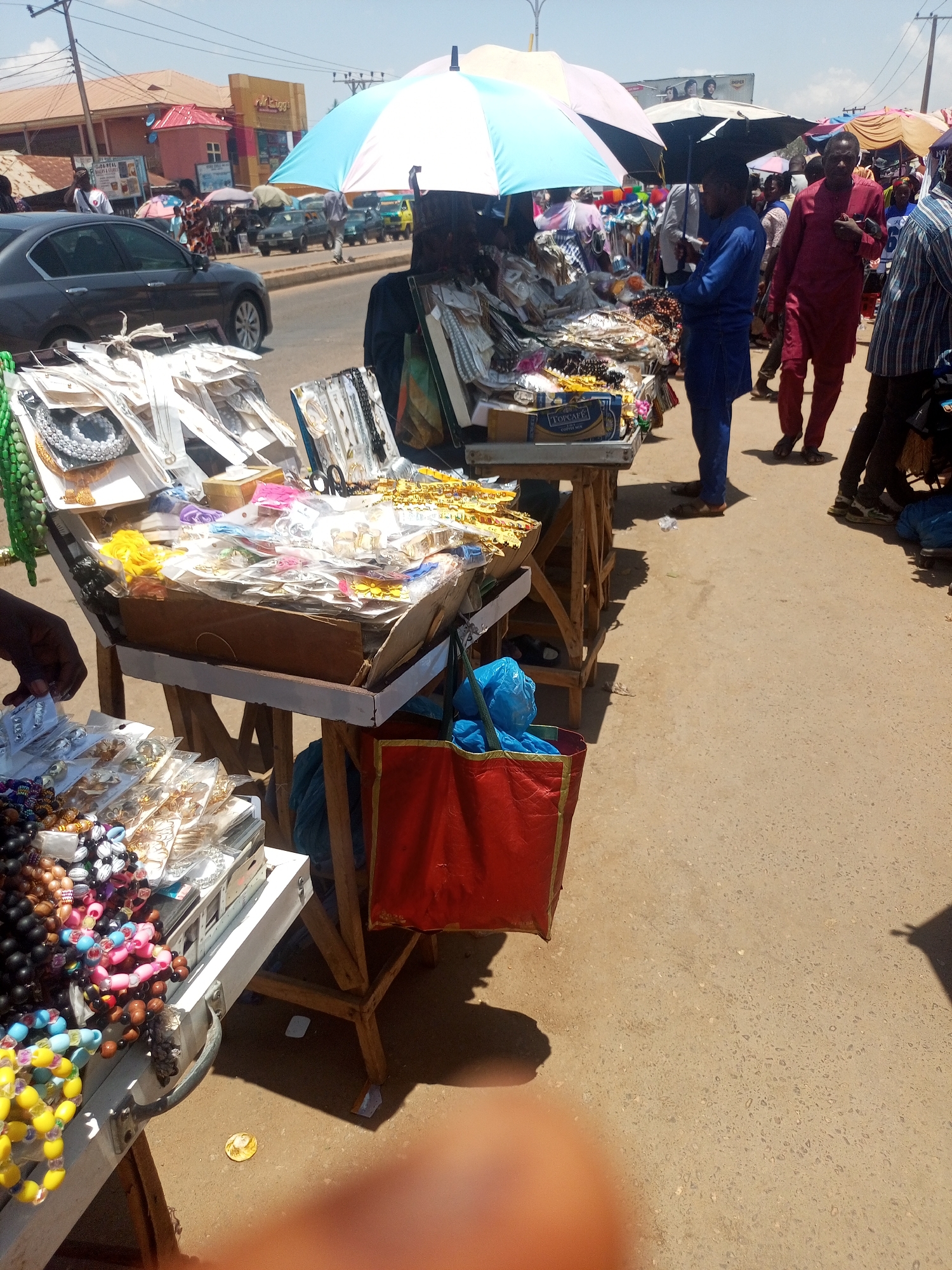 This image depicts an outdoor market scene in Sabon Tasha, Nigeria. Several vendor stalls line a paved area, displaying a variety of jewelry and accessories, including necklaces and bracelets, arranged on tables or in display cases. People, potentially customers or vendors, are standing around the stalls, some interacting with the merchandise. Most of the stalls are covered by parasols. In the background, there is a street with a black car, buildings, and traffic, under a bright sky. The ground is light-colored and textured. A partially visible hand appears in the lower part of the image.