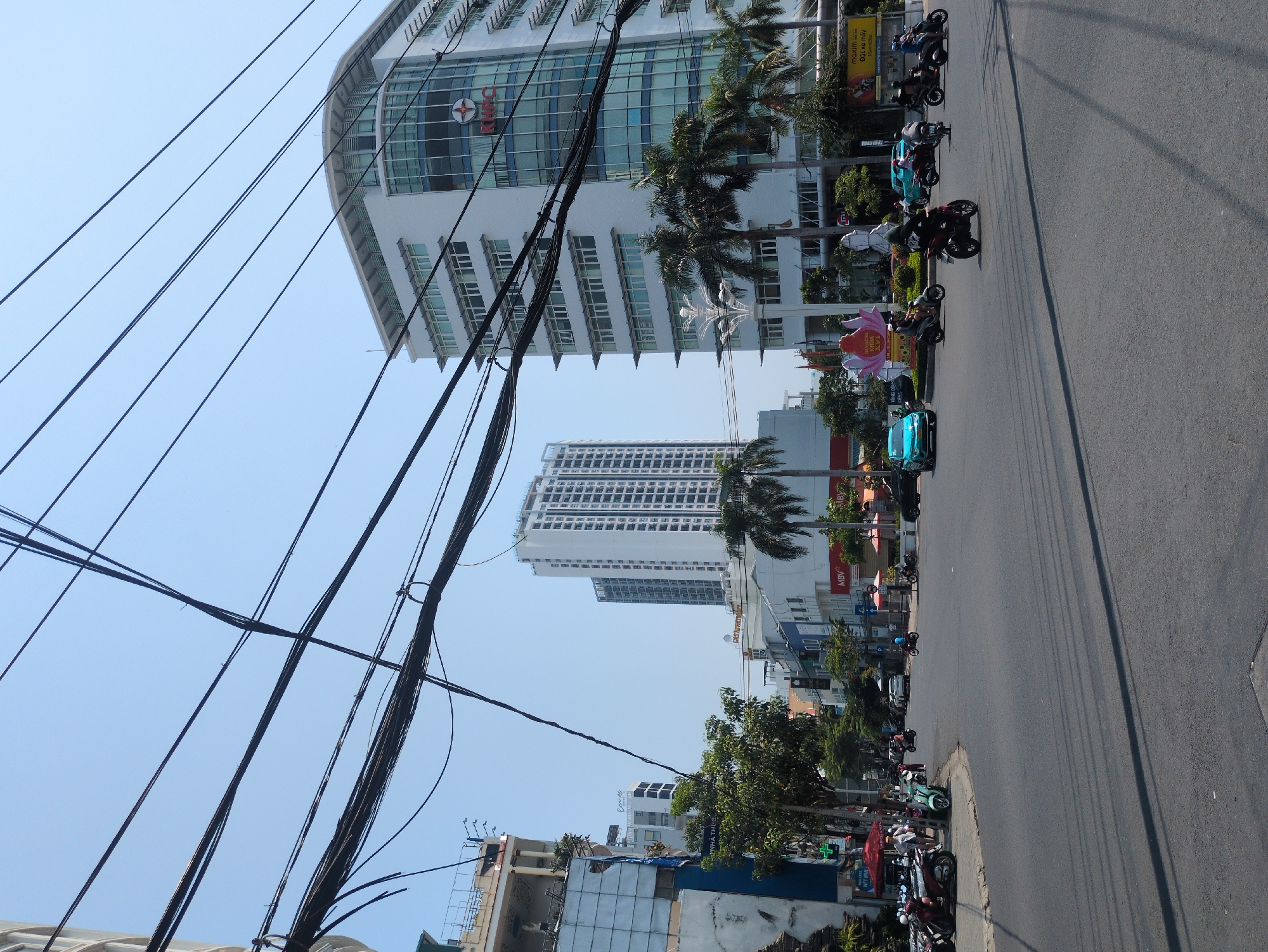 The image shows a street scene in Nha Trang, Vietnam, characterized by a street with scooters parked along the side. Buildings of varying heights line the street. Power lines crisscross the sky. The weather appears clear and sunny. Palm trees are interspersed along the street, adding to the tropical urban environment.