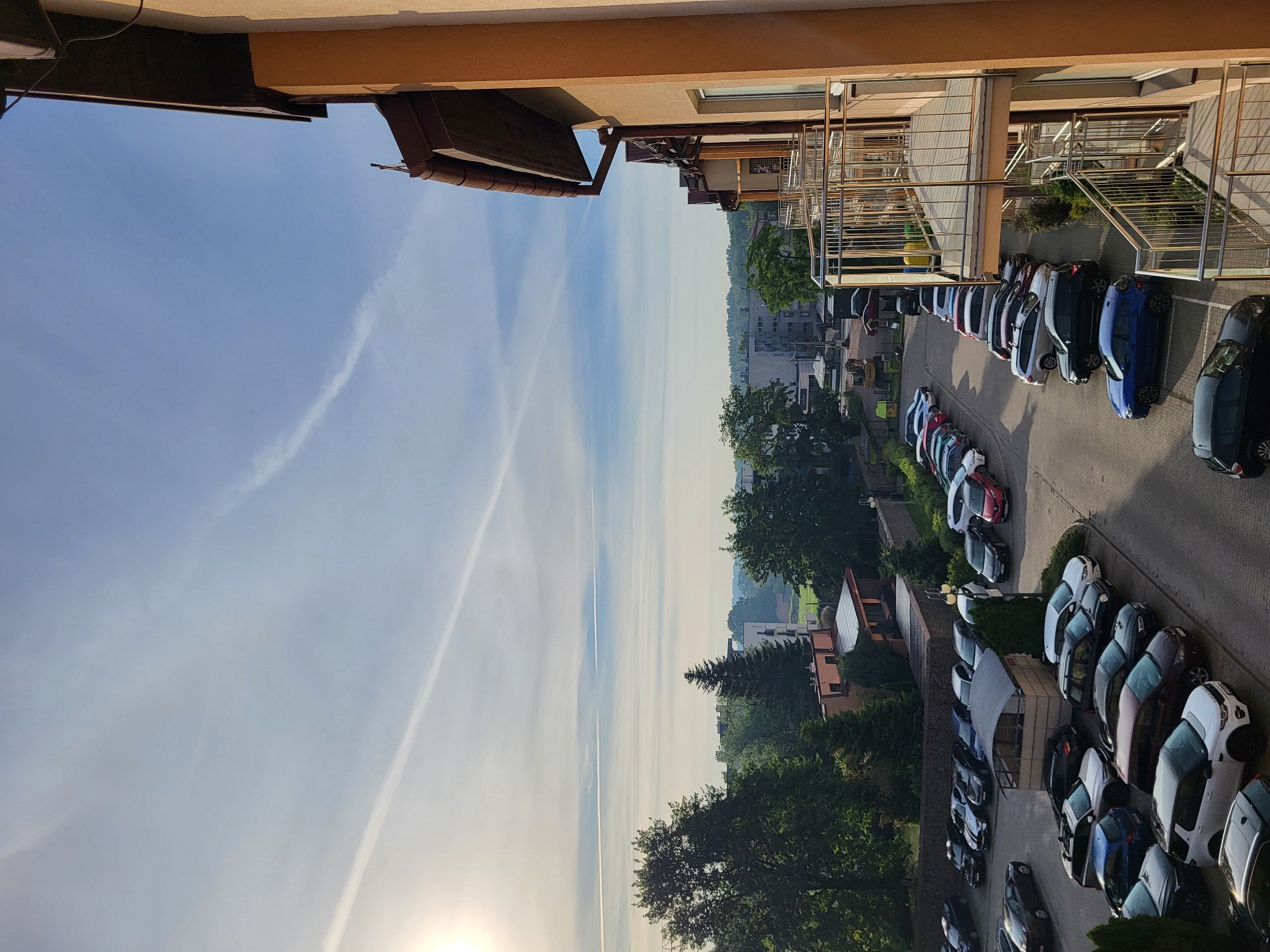 The image depicts an outdoor parking lot scene in a city. The parking lot is paved with interlocking bricks and is filled with various parked cars. The cars are of different makes and colors, including sedans, SUVs, and smaller vehicles. Some of the cars are parked in rows, while others are more scattered. In the background, there are buildings, trees, and a clear sky with some wispy clouds. The sun is visible in the upper left corner of the image, suggesting it is either early morning or late afternoon. The perspective is from a slightly elevated position, looking down on the parking lot.