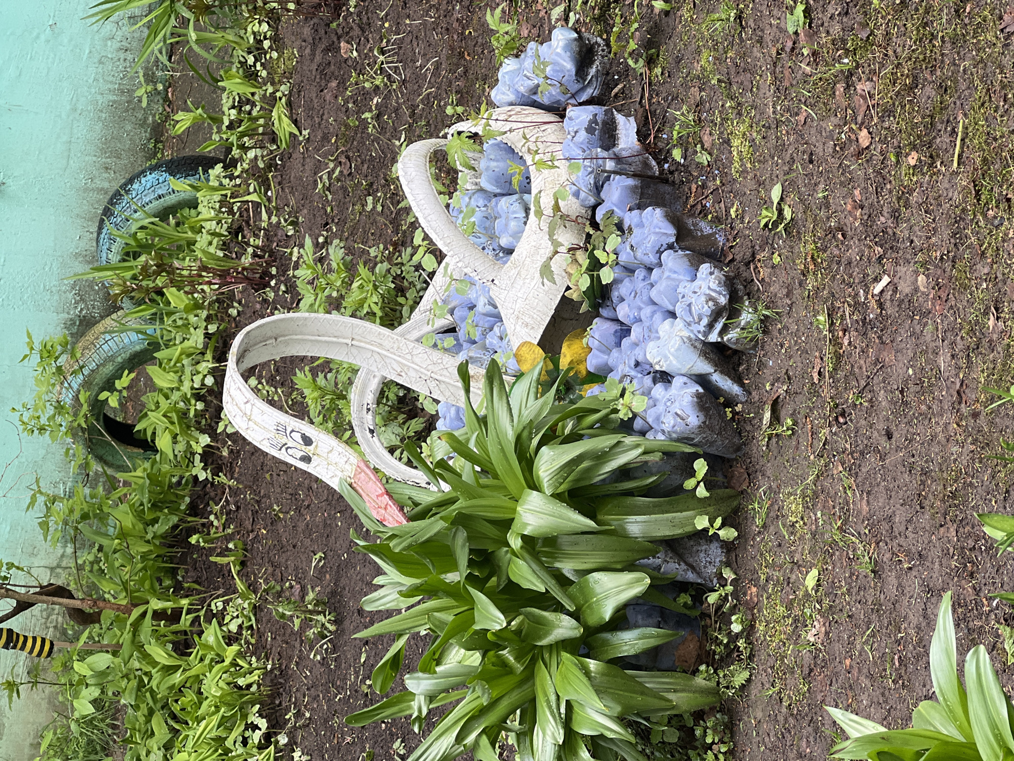 The image shows a garden scene in what is identified as the city of Chaikovsky, Russia. The central focus is a decorative swan constructed from repurposed car tires, painted white with cartoonish eyes and a pink beak. The swan appears to be part of a larger arrangement that resembles a flower basket, also made from tires and painted blue. Plants, including leafy green vegetation, surround and partially obscure the tire structure. Bare earth is visible in the background, along with some small green plants. A portion of a building or wall painted a light blue color is visible on the left side of the frame. Two blue-painted car tires are visible near the building.