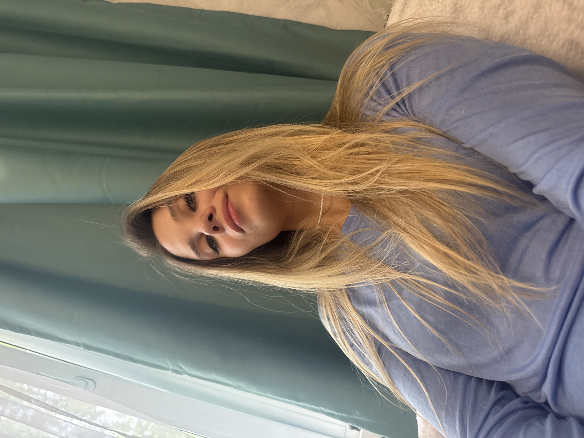 A young woman with long blonde hair is looking up at the camera, her head tilted slightly. She is wearing a light blue long-sleeved shirt and a delicate white beaded necklace. The background consists of a window with sheer white curtains on the left and a teal curtain on the right. A portion of a white textured surface is visible on the far right. The lighting suggests an indoor setting, likely daytime, with natural light coming from the window. The woman's expression is serene, with a hint of a smile. The image appears to be a selfie taken from a low angle.