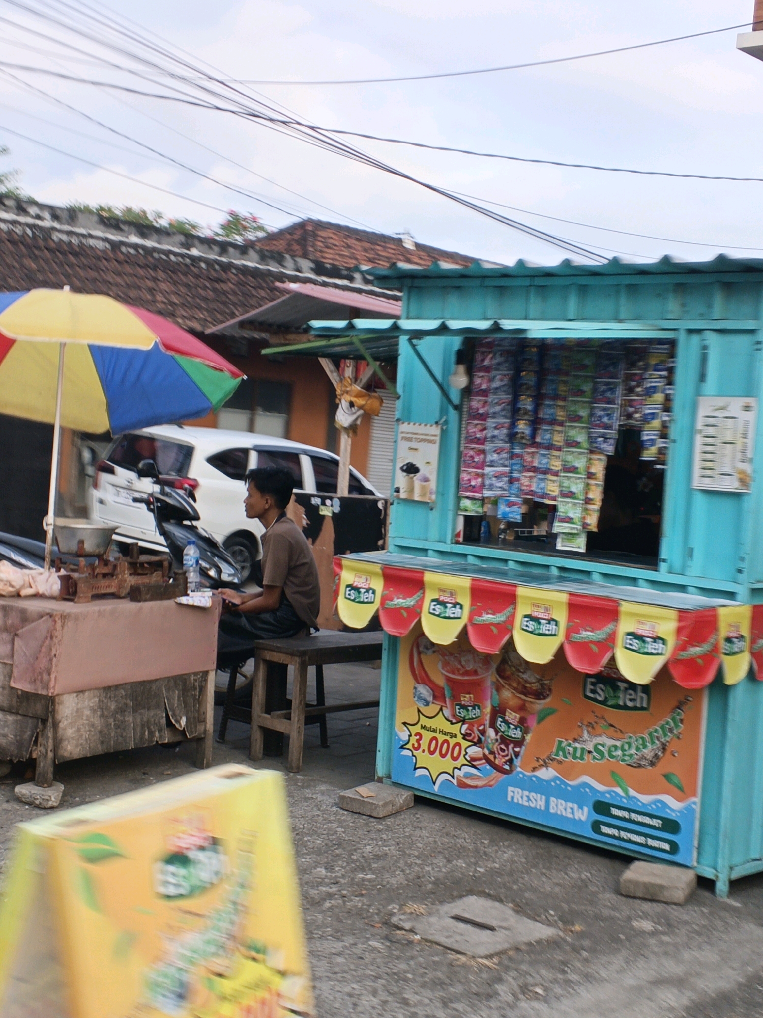 A young man with short, dark hair sits on a wooden stool beside a street stall in Sukawati, Indonesia. He is wearing a brown t-shirt and dark pants. The stall is a bright turquoise blue container, with a window displaying an assortment of packaged beverages. Below the window, a colorful banner advertises "Es Teh" (iced tea) with images of iced tea drinks and the text "Ku Segarrr" and "FRESH BREW". A price of "3.000" is also visible. To the left of the stall, a red, brown, and pink umbrella is partially visible. In the background, a white van is parked on the street, and the roofs of houses with terracotta tiles are visible. Overhead, numerous electrical wires crisscross the sky. In the foreground, a blurry yellow sign with graphics is visible, partially obscuring the lower portion of the scene. The ground is paved with asphalt.