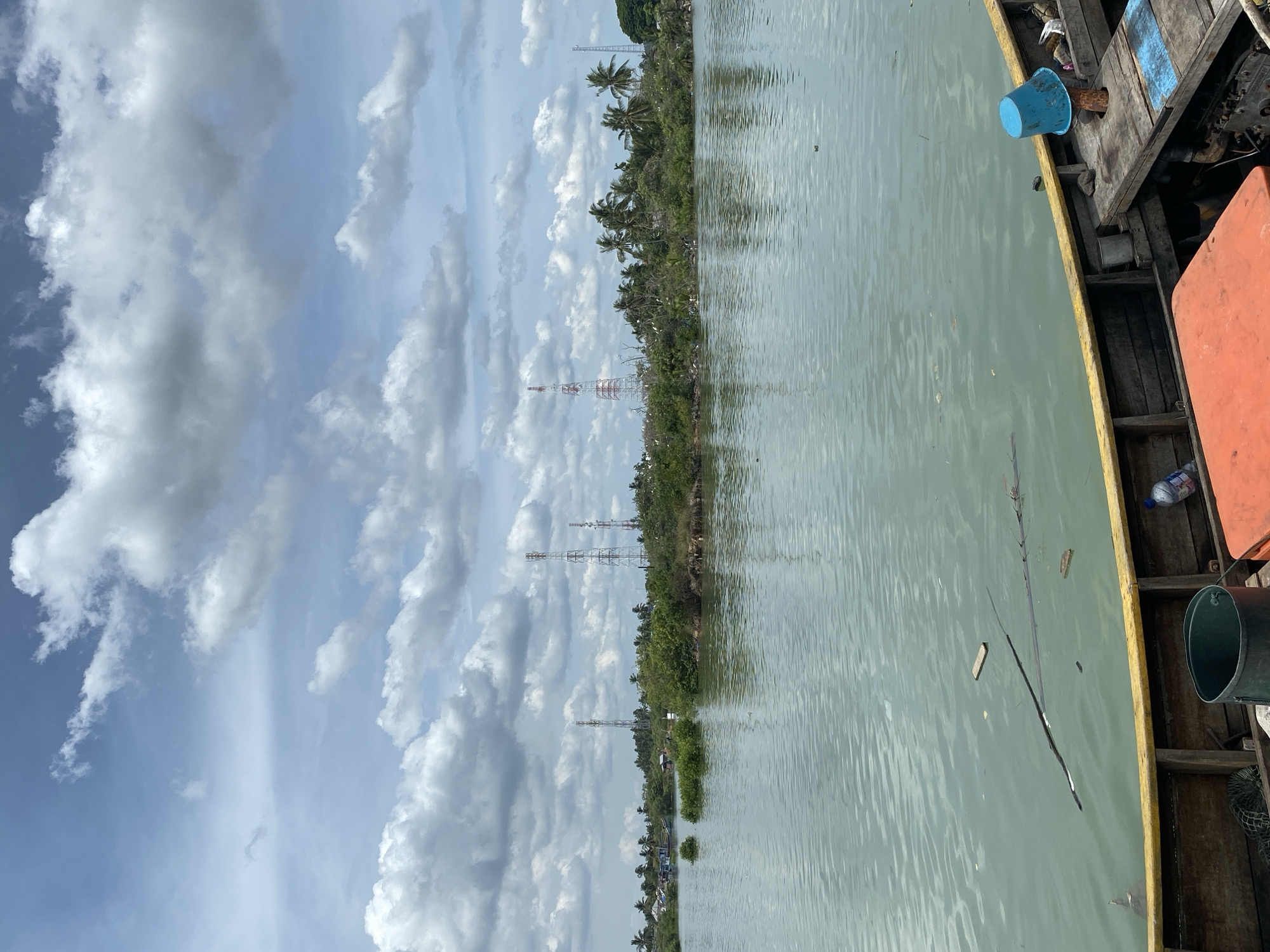 The image depicts a waterway in Tanjongkapai, Indonesia, with a partially visible wooden boat in the foreground, on the right. The boat contains various items including a blue bucket, an orange object, a metal bucket, and a plastic bottle. To the left of the boat is water and beyond the water is a land mass covered in trees, including palms, and vegetation along the waterfront. There are four telecommunications towers erected on the landscape. The sky above has light blue patches and is filled with white, billowy clouds.