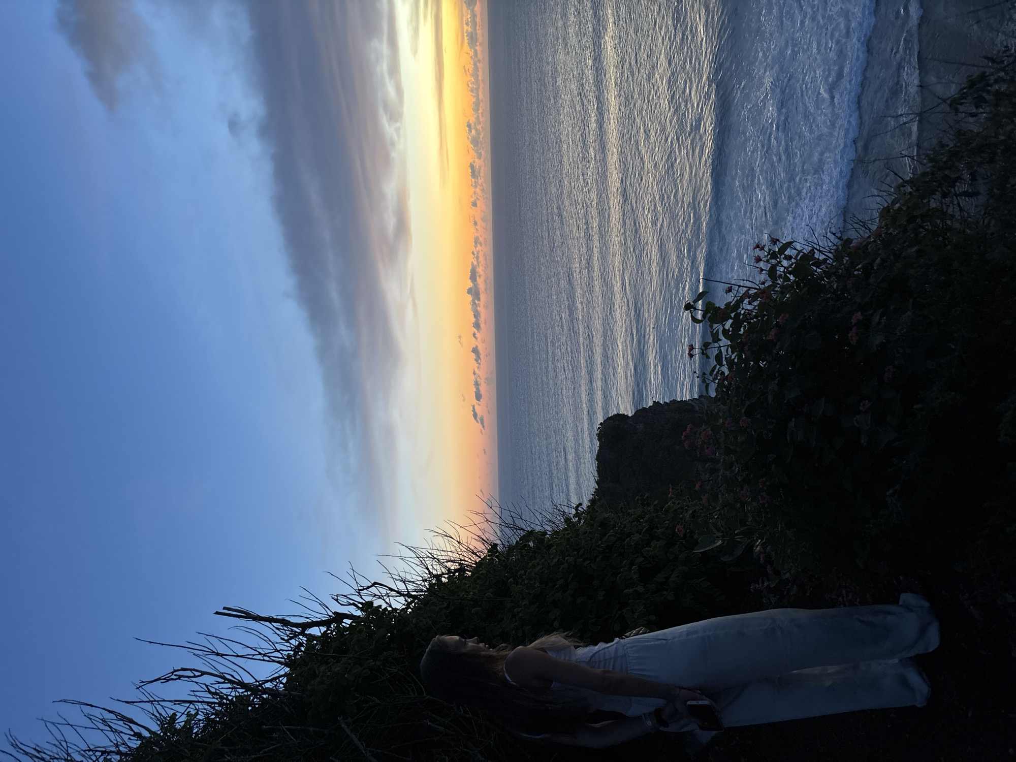 A woman lies on a cliff overlooking the ocean at Uluwatu, Indonesia. She is dressed in light-colored clothing and is positioned among the greenery on the cliff's edge. The sky displays hues of blue and orange, indicating a sunset or sunrise. The ocean stretches out in front of her, with visible waves. The scene is mostly in shadow, suggesting either early morning or late evening.