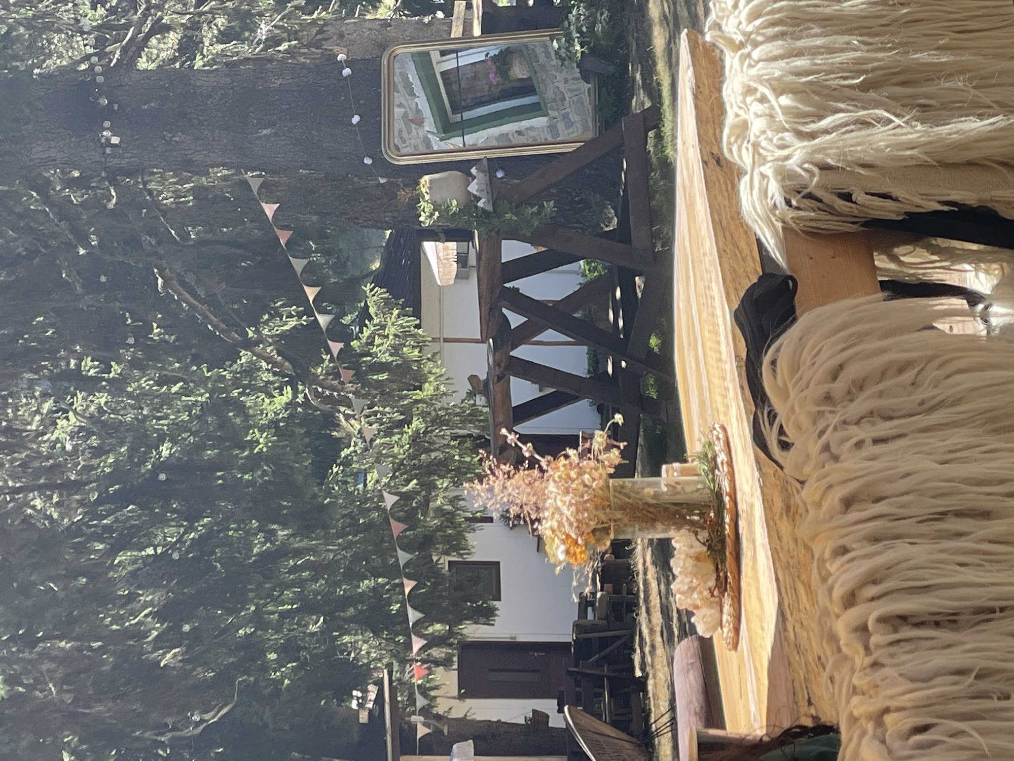 This image depicts an outdoor setting, likely a cafe or garden area, with a rustic and bohemian aesthetic. In the foreground, a wooden table is visible, draped with long, shaggy cream-colored fabric, possibly a blanket or rug. A glass vase holding dried flowers and a decorative element of white crystals sits on the table.
Behind the table, a large tree trunk is prominent, adorned with string lights and a triangular pennant banner in muted colors. A large, ornate gold-framed mirror leans against the tree, reflecting a stone building with a green-trimmed window and flowers. More pennant banners are strung between trees in the background, with a white building with a dark door visible behind them. The overall environment suggests a relaxed, natural ambiance, possibly in a park or forest setting. The specific location in Smolyan, Bulgaria is not discernible from the image itself.