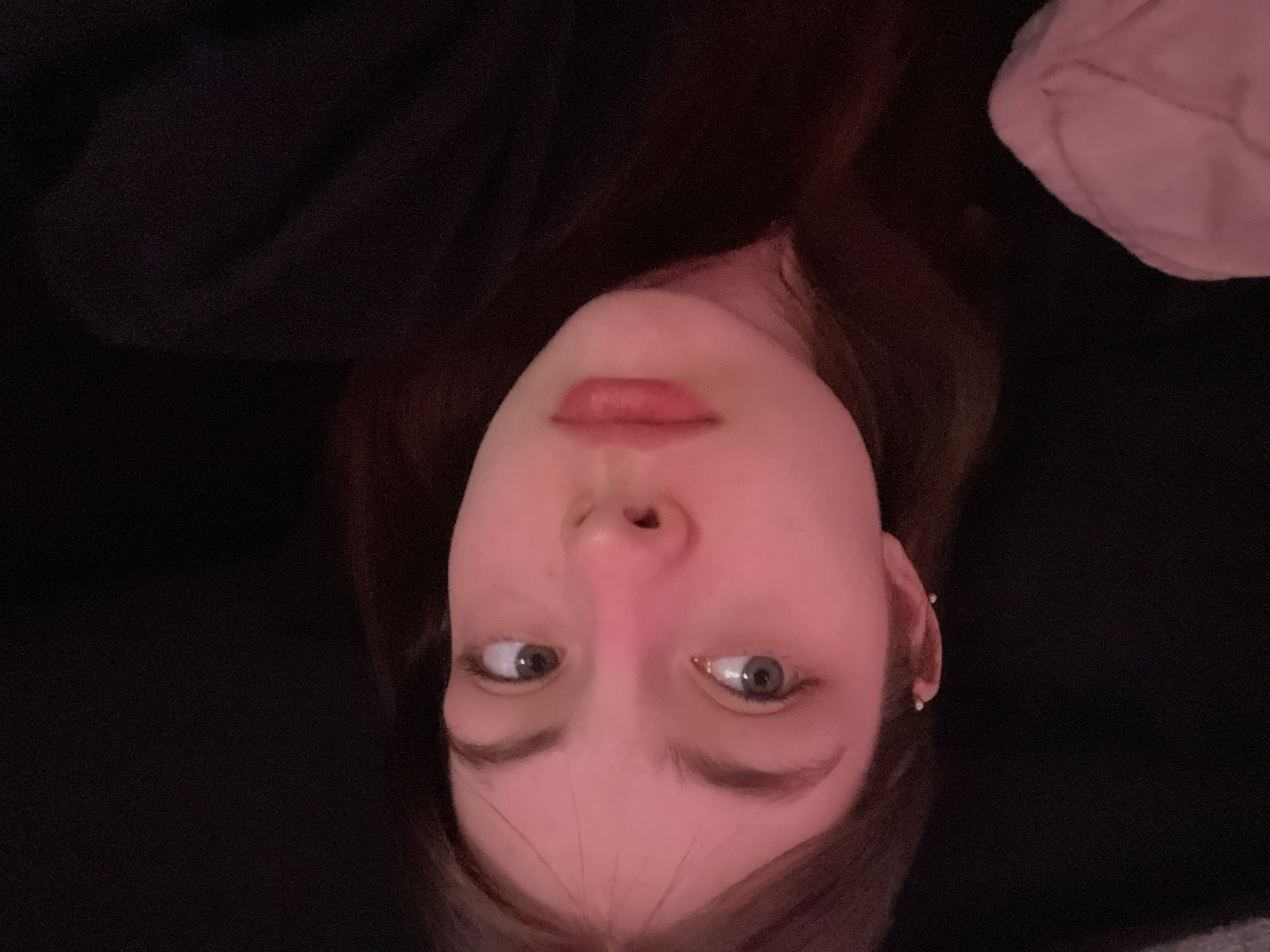 This close-up portrait, taken in Saint Petersburg, Russia, features a young person, likely female, positioned upside down and centrally in the frame. Her face is illuminated by a soft, pinkish light, which highlights her fair complexion, light-colored eyes, and full lips adorned with reddish-pink lipstick. Her gaze is directed to her right (viewer's left), conveying a thoughtful or quizzical expression. Dark brown hair frames her face, and a small, stud earring is visible in her right ear.
The background is predominantly dark and indistinct, suggesting a dimly lit indoor environment, possibly a room at night. Hints of light pink, soft fabric (perhaps a blanket or garment) are visible in the upper right corner, and dark clothing (like a sweater or hoodie) can be seen on the upper left, reinforcing an intimate and relaxed setting. The inverted orientation of the image provides an unconventional and artistic perspective, characteristic of a casual selfie. No text is visible within the image.