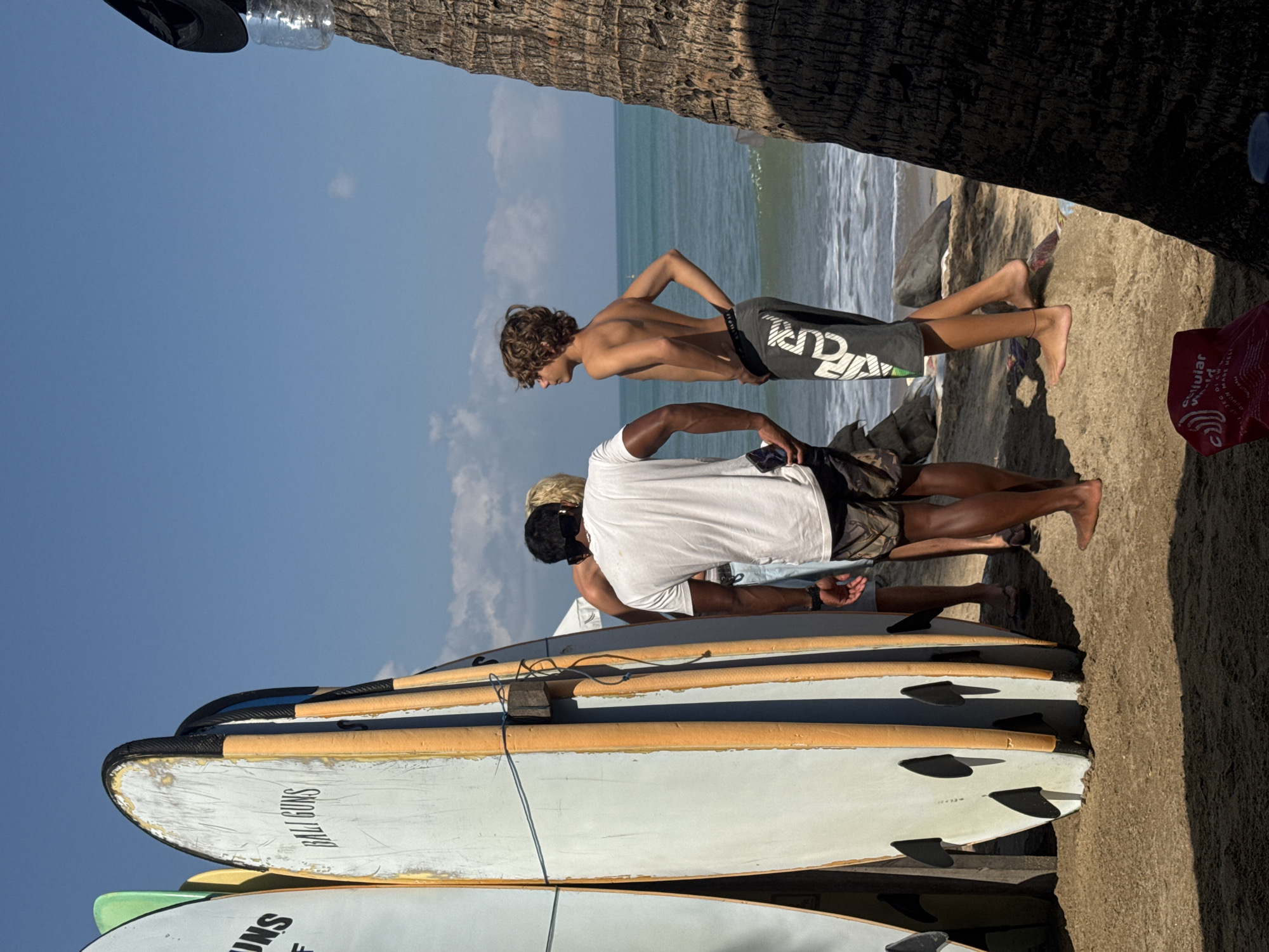 The image depicts a sunny day on a beach in Kuta, Indonesia. Several surfboards are leaning against each other, with the brand "BALI GUNS" visible on one. Three young men are present. One man, wearing a white t-shirt and patterned shorts, is facing away from the camera, holding a phone. Another young man, shirtless and wearing dark shorts with white lettering, is looking out at the ocean. A third man, partially visible and wearing a white t-shirt and light-colored shorts, stands between the two. The ocean is calm with small waves, and the sandy beach is sparsely populated. A large palm tree trunk is in the foreground on the right side of the image, casting a shadow. The sky is clear blue with a few wispy clouds.