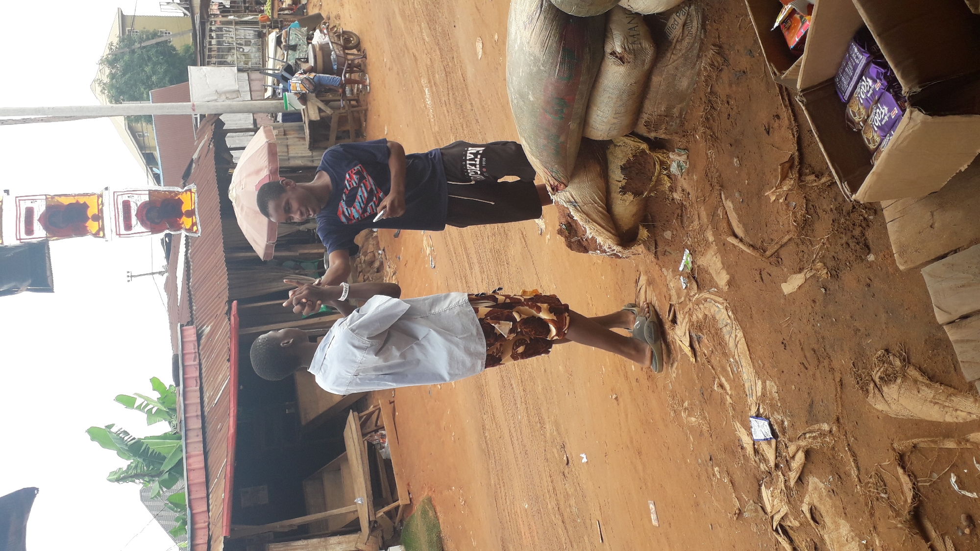 The image depicts an outdoor scene in Nkpor, Nigeria. Two men are engaged in a conversation. One man is wearing a light-colored shirt and patterned shorts, while the other is wearing a dark blue t-shirt with a white design and dark shorts. The ground is dirt. To the right, there are sacks of goods and boxes of packaged food. In the background, there are buildings with corrugated metal roofs. There's an umbrella and some plants. A man is seated on a wheeled cart further down the road. There's a sign hanging above.