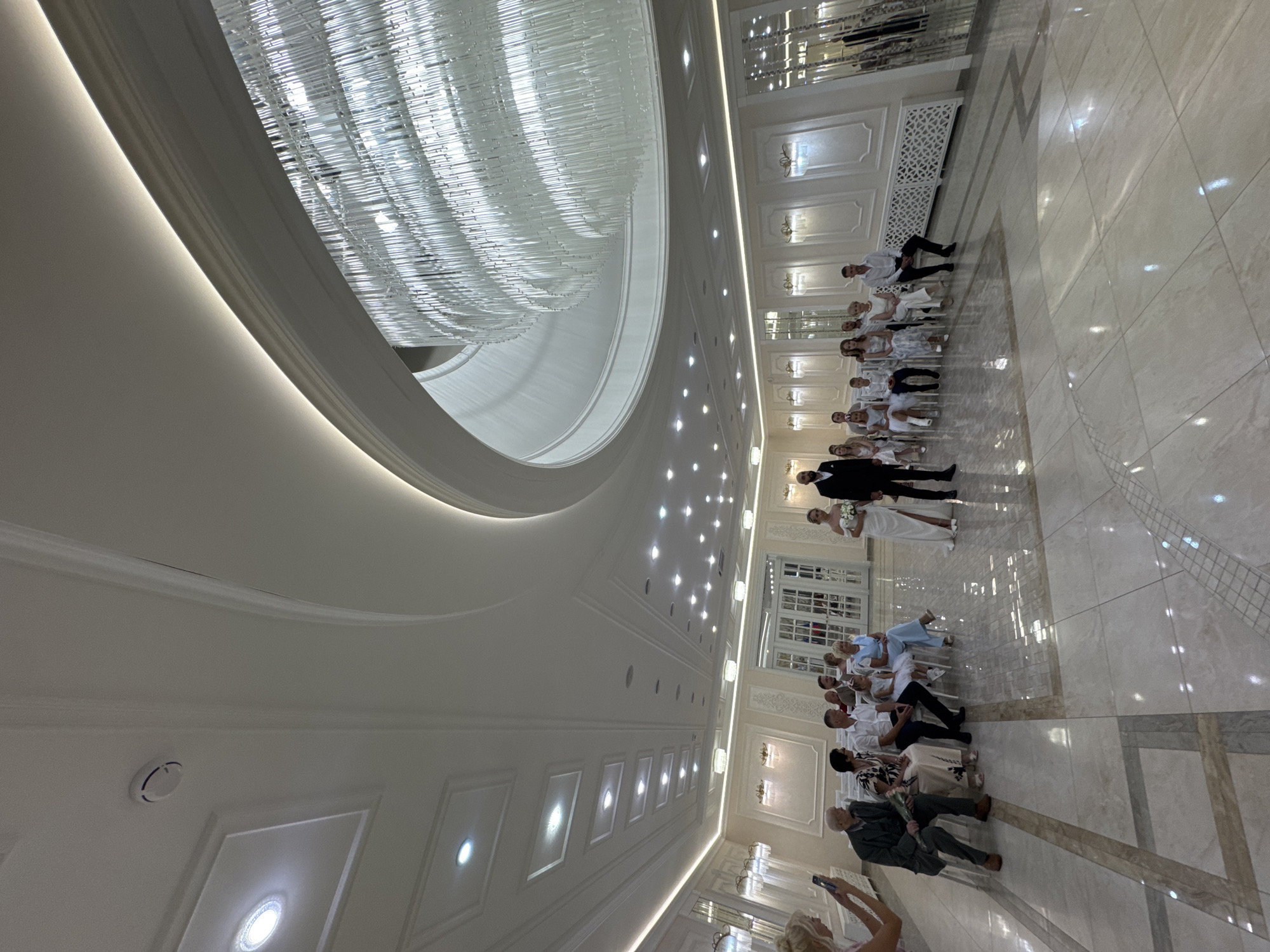 This is a wide-angle, eye-level, full interior shot of a wedding ceremony taking place in a spacious, modern hall. The hall features a high, intricately coffered ceiling with recessed lighting and a large, multi-tiered, glass-rod chandelier. The walls are white and adorned with decorative paneling and sconce lighting. The floor is polished white tile with darker inlaid geometric patterns.
In the center of the hall, a bride in a white gown and a groom in a dark suit stand facing an audience. The bride holds a bouquet. The groom stands to her right.
To the left of the bride and groom, a group of guests are seated on chairs arranged in rows. On the far left, a woman in a pink top is taking a picture with her phone. Further into the hall, seated guests are dressed in various casual and semi-formal attire. Among the seated guests are individuals of different ages, including men, women, and children. Some of the children are dressed in white.
To the right of the bride and groom, more guests are seated, also in rows. These guests are similarly dressed. The image captures a formal event, likely a wedding ceremony, in a well-lit and elegant indoor venue.
The provided information states the location is in Tolyatti, Russia. Without additional context or specific landmarks within the image, it is impossible to pinpoint the exact location within Tolyatti. However, the visual elements suggest a banquet hall or a dedicated event space suitable for ceremonies.