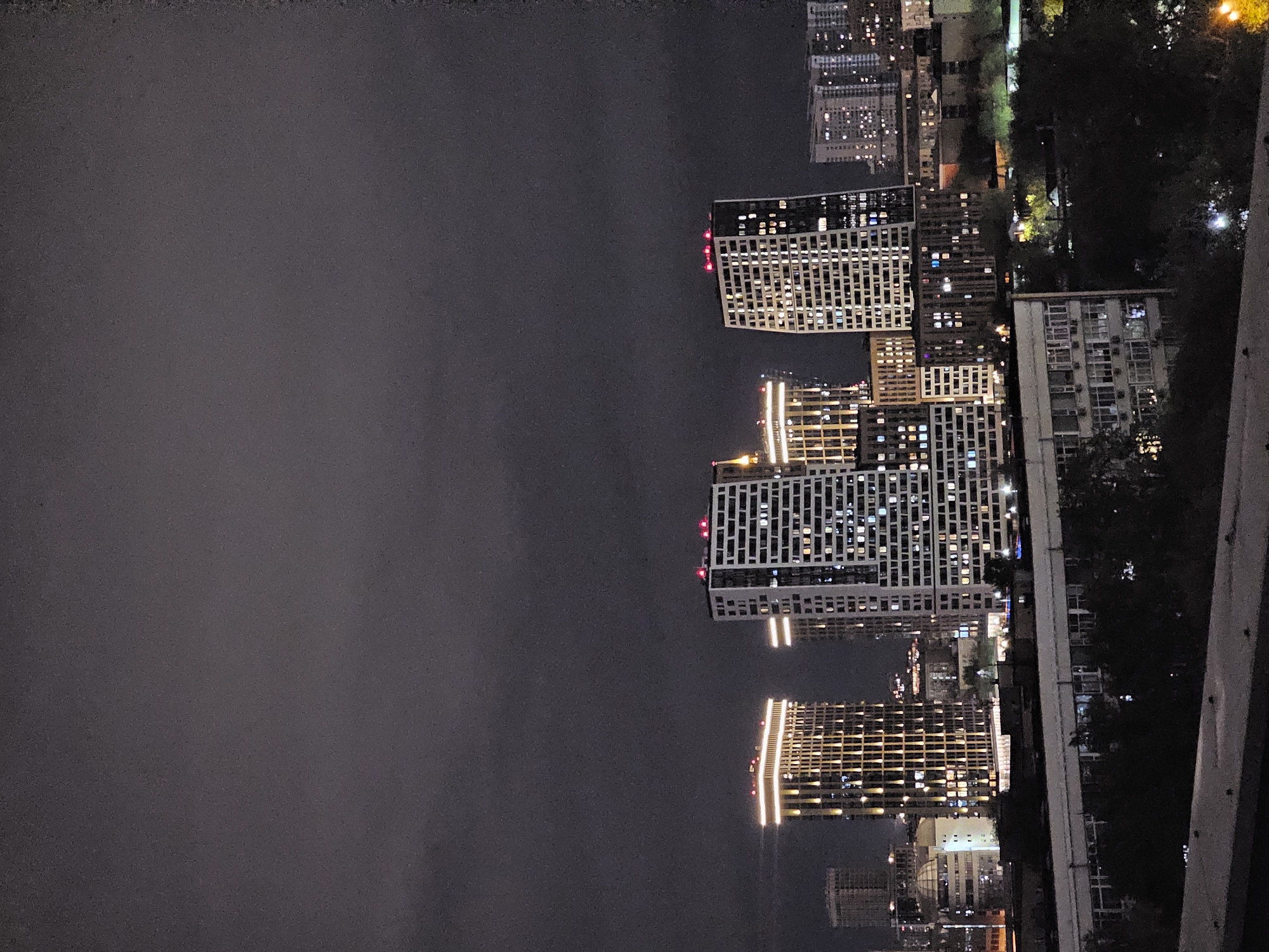 The image captures an urban cityscape at night, likely in Moscow, Russia, based on the architectural style of the buildings. The focus is on a cluster of modern high-rise buildings with lit windows, indicating residential or commercial occupancy. The buildings vary in design, with some having a grid-like façade and others appearing more uniformly lit. The sky is dark and overcast. The shot is taken from an elevated position, possibly a balcony or rooftop, and a railing is visible. The buildings are lit with orange and white lights. Green foliage is present and interspersed between the buildings.