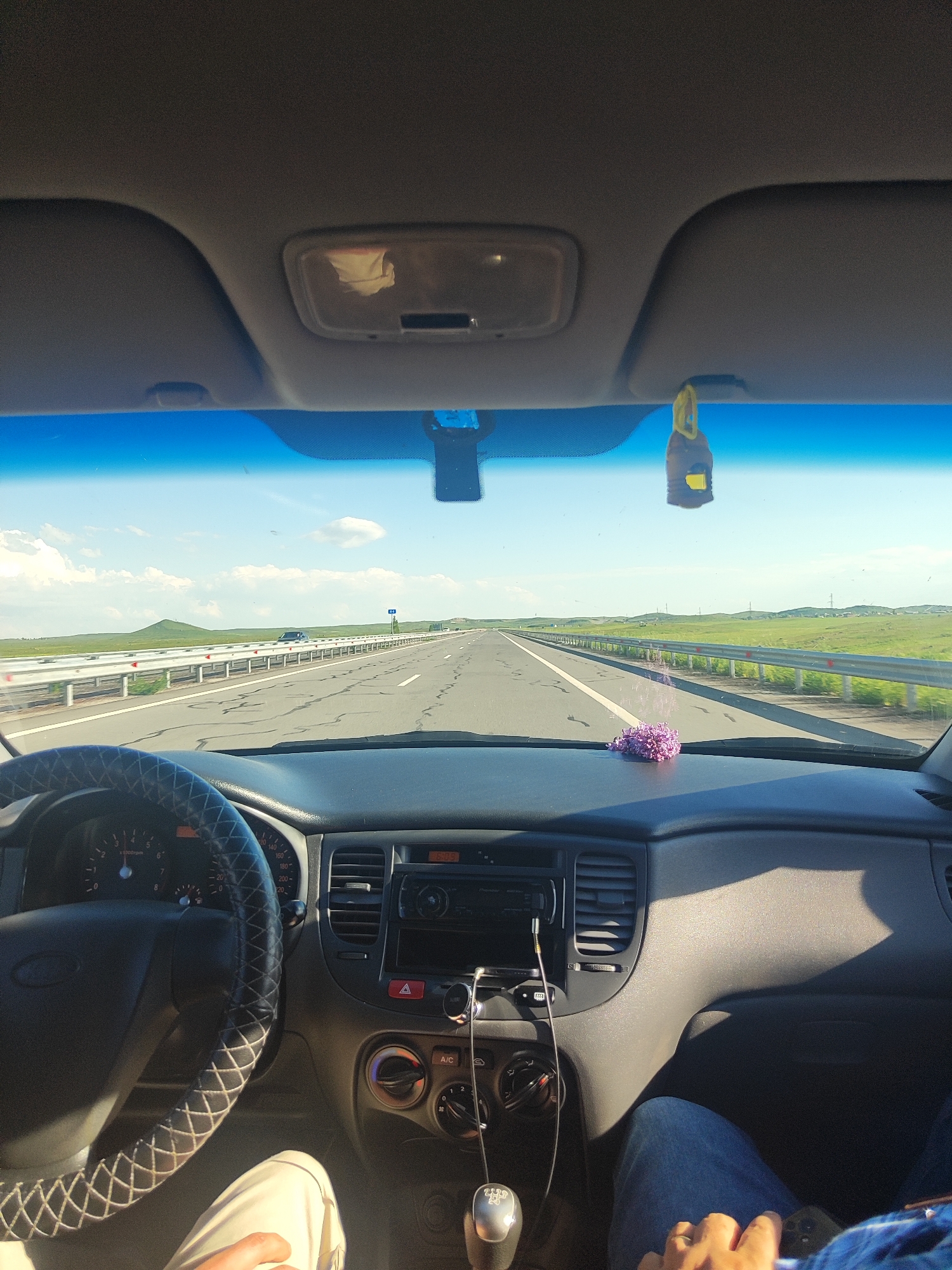 The image is taken from the interior of a car driving on a highway. The perspective is from the driver's seat, looking forward. In the foreground, the steering wheel and dashboard are visible. The steering wheel is covered in black material with a white cross-stitch pattern. The dashboard includes the instrument cluster, air vents, and a car stereo. A small bunch of purple flowers is placed on the dashboard.
Outside the car, a multi-lane highway stretches into the distance under a bright, blue sky with scattered white clouds. The landscape on either side of the highway is grassy and rolling, with a few distant hills. Guardrails line the sides of the road. The sun appears to be high in the sky, suggesting it is daytime, likely midday or afternoon. The weather seems clear and sunny.
A yellow air freshener hangs from the rearview mirror. The reflection in the mirror shows a white object, possibly a tissue. The scene appears to be a road trip through a relatively open and natural landscape. There are no explicit emotional cues, but the image conveys a sense of travel and journey. No visible text is present other than indicators on the car's dashboard.