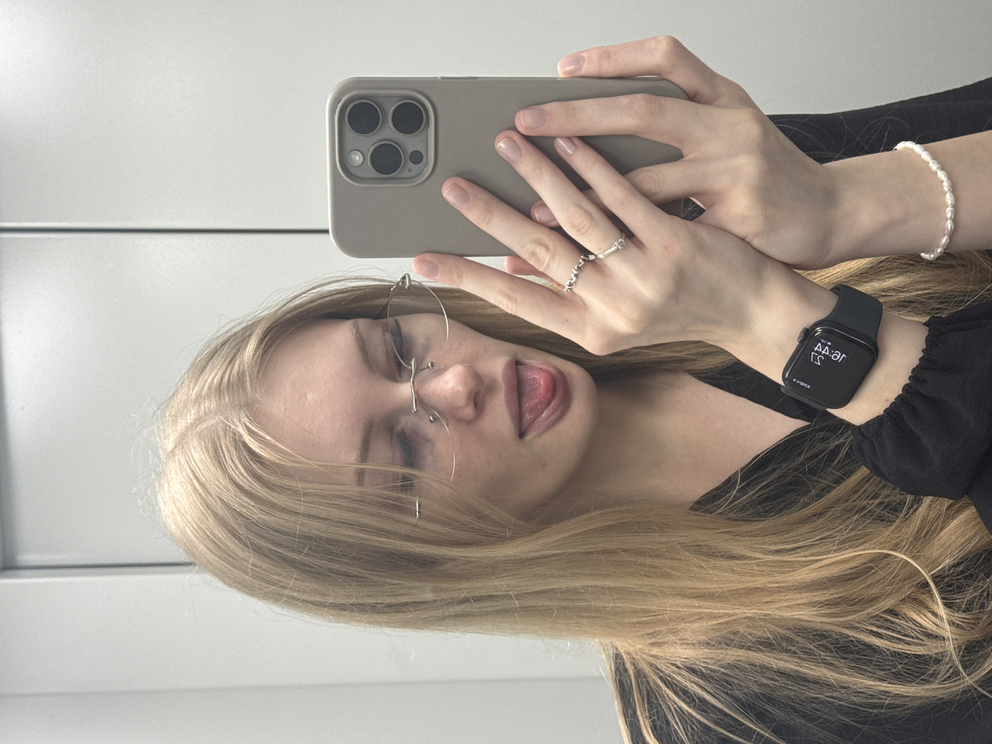 A young Caucasian woman with long blonde hair and glasses is taking a selfie in a grey, minimalist room. She is wearing a black top and a smartwatch on her left wrist. She is sticking her tongue out slightly at the camera. She is holding a grey iPhone in her right hand, with her left hand resting on the phone. The room appears to be clean and modern, with smooth, light grey walls. There is a vertical seam in the wall behind her, suggesting it might be a door or a panel. There are no other people or objects visible in the frame. The overall impression is of a casual, personal moment captured in a neutral environment. The image does not contain any information that can be used to determine the location in the city of Perm, Russia.
