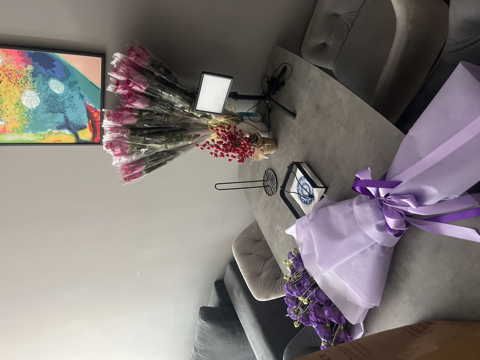 The image depicts a still life composition arranged on a grey stone table, likely in a modern residential or office setting. In the foreground, a large bouquet of purple flowers is elegantly wrapped in light purple and white paper, secured with a satin ribbon. To the left, partially visible, is a brown paper bag. Further back on the table, a smaller arrangement of red berries in a burlap pot adds a touch of color. To the right, a small LED light on a tripod stands prominently, suggesting preparation for photography or videography. Behind this, a bunch of pink roses, also wrapped in clear plastic, are arranged. A black metal stand holding napkins and a blue-patterned object rests near the purple flowers. In the background, a dark grey sectional sofa is visible, and above it hangs a colorful abstract painting. The overall ambiance suggests a space dedicated to aesthetics or creative endeavors. The location is not specified in the image.