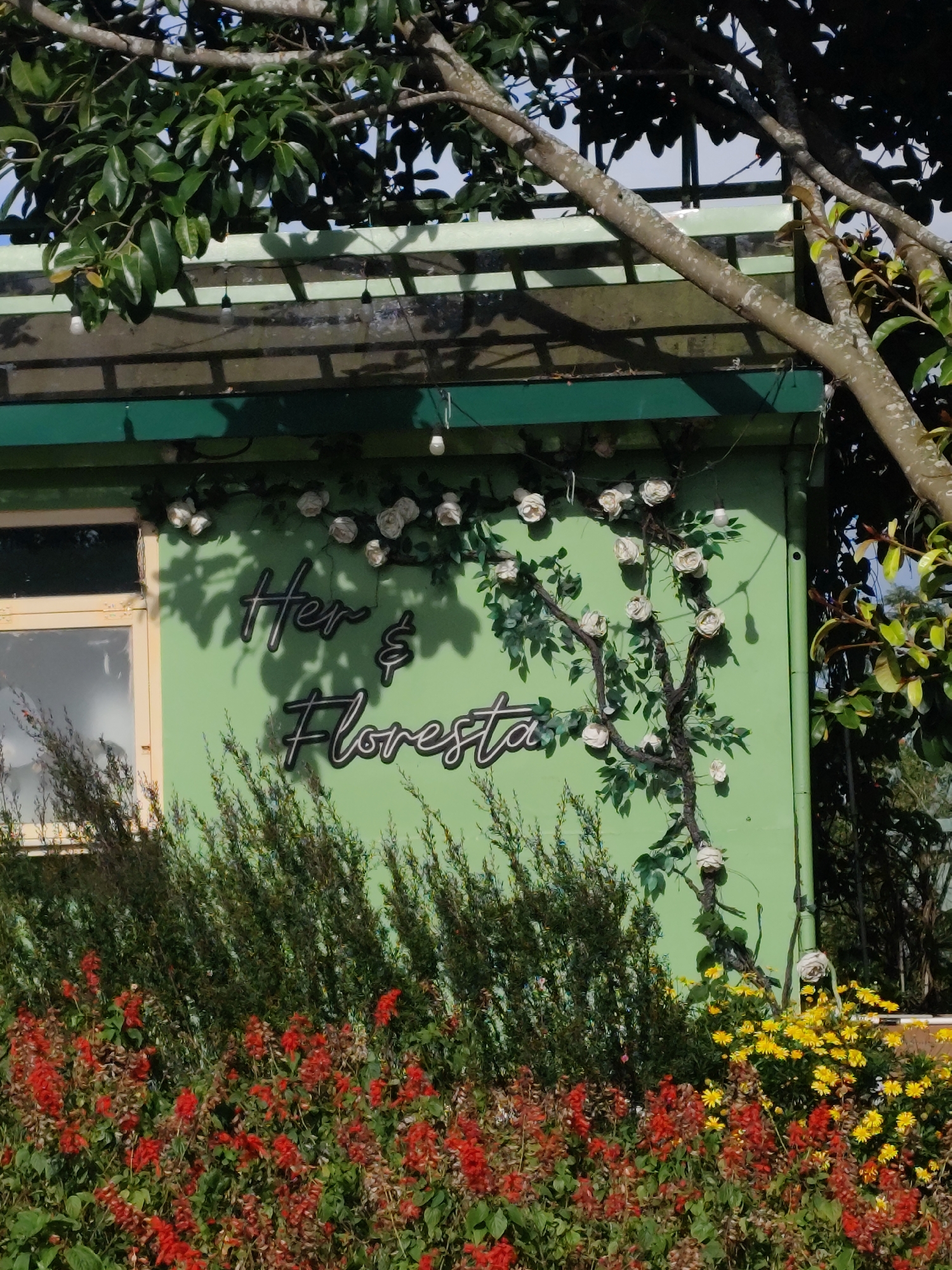 A light green building with a light green awning is visible, partially obscured by tree branches and foliage. The building has a window with a light tan frame. Text in black with a white outline reads "Her & Floresta" painted on the wall, accompanied by a dark vine with white roses. In front of the building, a dense arrangement of red and yellow flowers blooms, with dark green bushes behind them. The scene appears to be outdoors, under natural lighting. This image was captured in Da Lat, Vietnam, a city known for its cool climate and abundant flower gardens, often referred to as the "City of Eternal Spring."
