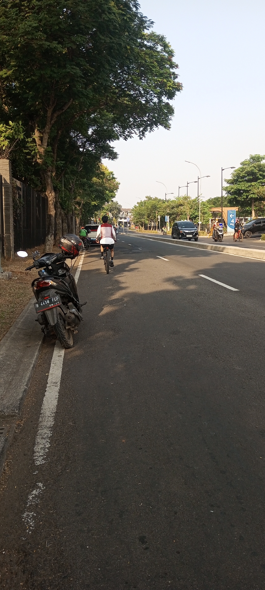 The image shows a street scene in Lambangjaya, Indonesia, during daylight. In the foreground, a parked black motorcycle is visible, facing left, with its license plate reading "B 4458 FCF 09-27". A person wearing a white shirt with red trim is cycling away from the camera on the right side of the road, which is paved with asphalt and has a white line marking the lane. Other vehicles, including cars and motorcycles, are present further down the road. The street is lined with trees on the left, and a fence is visible behind the trees. The sky is clear and bright, suggesting it is a sunny day. The overall environment appears to be a suburban or urban area with well-maintained roads and vegetation.