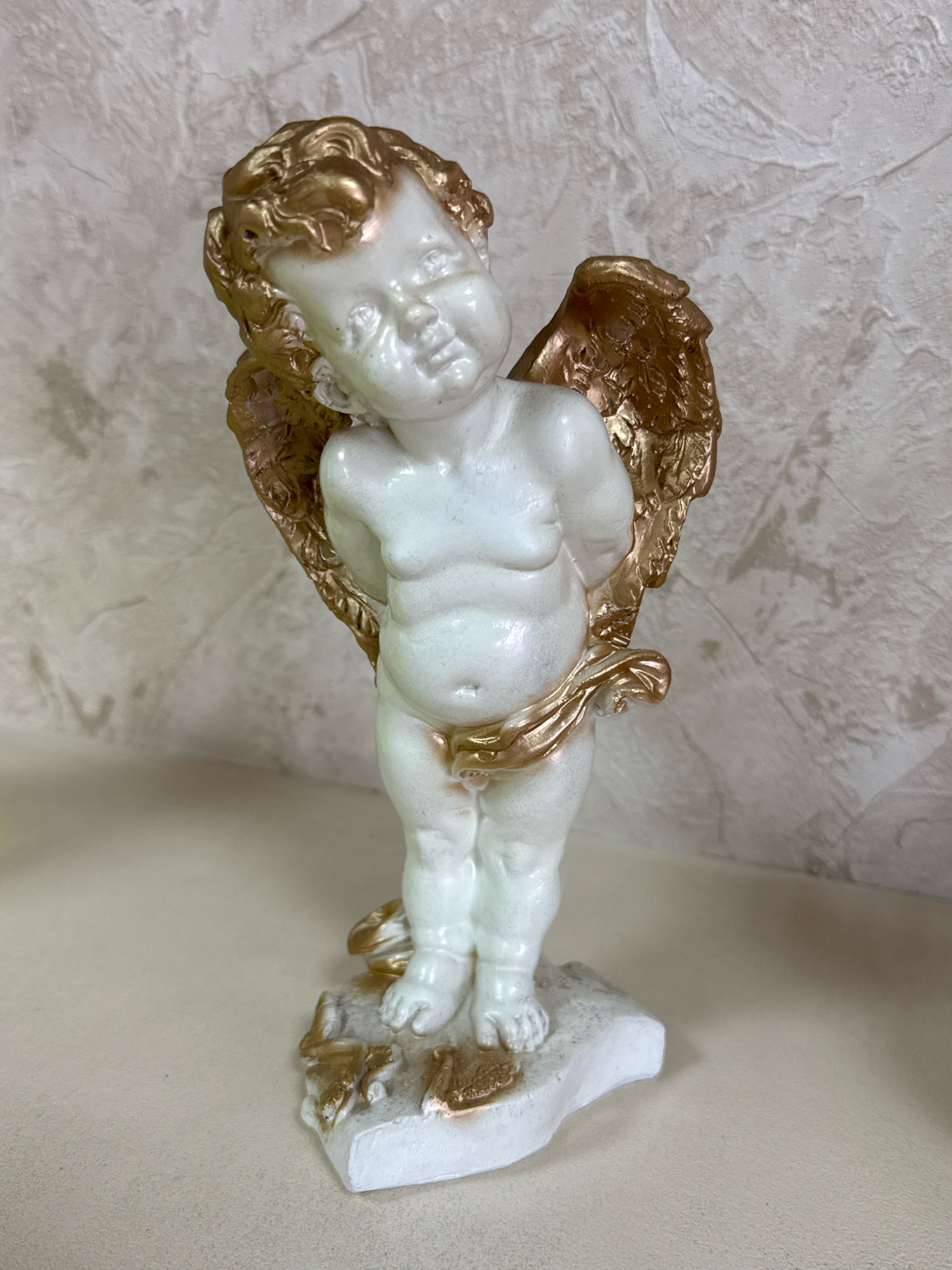 An ornamental cherub statue stands on a flat surface, its white body accented with gold details on its hair and wings. The statue is positioned against a textured, light-colored wall. The background appears to be an interior wall in a domestic setting, possibly in the village of Ryabovo, Russia, given the provided context. The lighting is even, suggesting a well-lit room. There are no people or animals in the image.