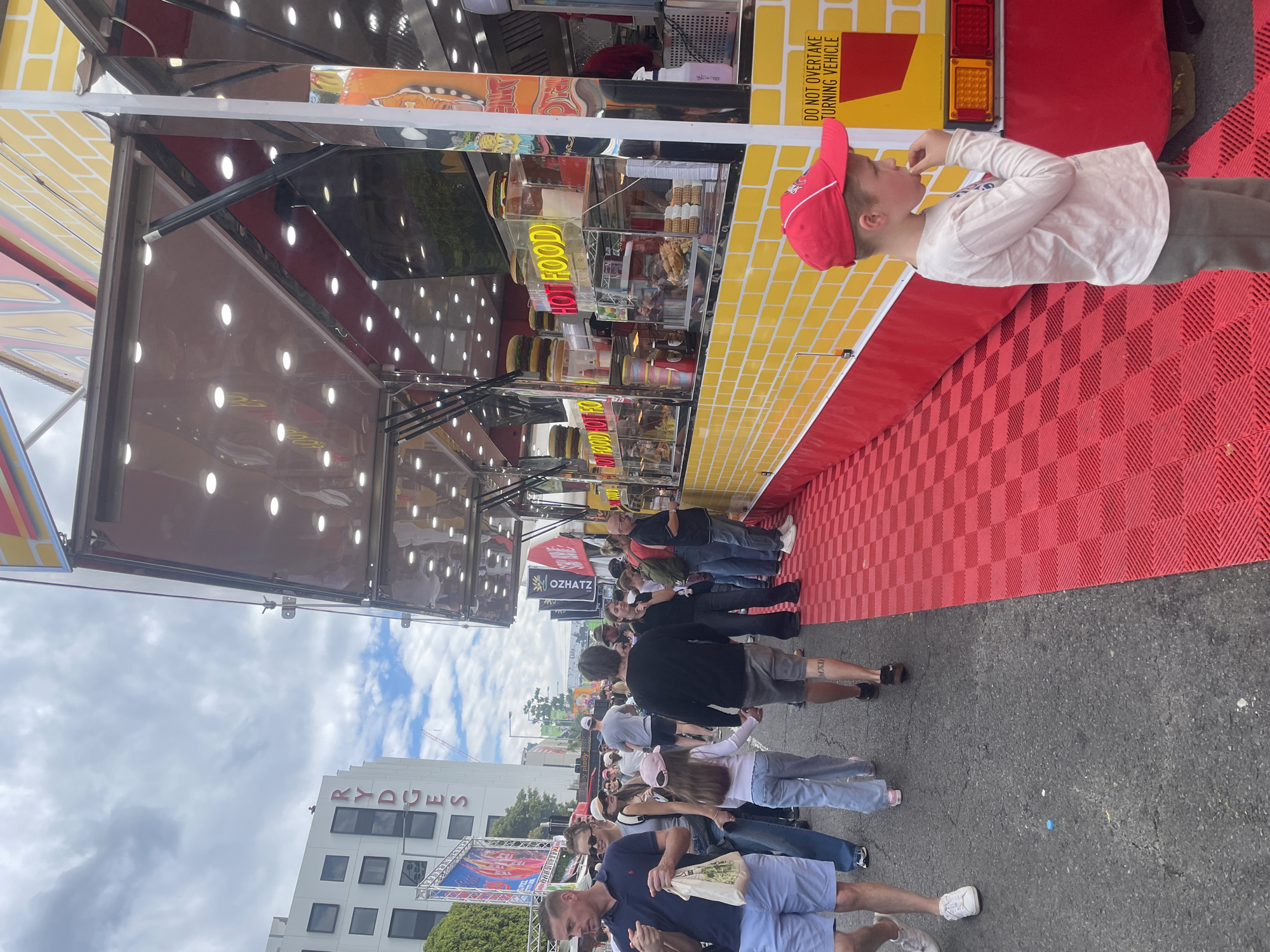 In Bowen Hills, Australia, a bustling street scene unfolds with a vibrant food truck as the central focus. The truck, painted with a bright yellow brick pattern and adorned with red trim, displays illuminated signs advertising "HOT FOOD." Inside the truck, rows of burgers and other food items are visible behind glass enclosures. A young boy in a red baseball cap stands in the foreground, looking towards the food truck and eating. He is wearing a long-sleeved shirt and pants.
Crowds of people mill about on a red checkered floor mat that extends along the sidewalk in front of the food truck. A sign on the truck reads "DO NOT OVERTAKE TURNING VEHICLE." In the background, a modern white building with dark square windows is visible, with the word "RYDGES" prominently displayed in red letters on its facade. The sky is overcast with scattered clouds. The scene captures the lively atmosphere of an outdoor event or market.