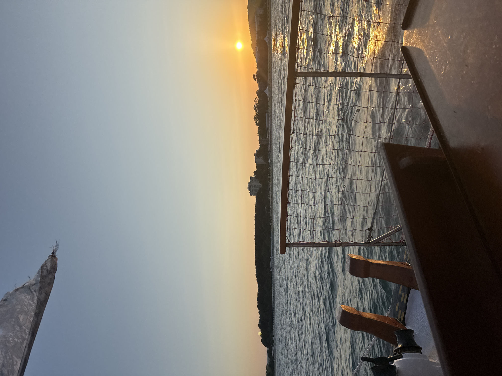 The image depicts a serene sunset over the Black Sea, as viewed from the deck of a boat. The sun, a bright orb of orange, is low on the horizon, casting a golden glow across the water. The sky transitions from a soft blue near the top to a warm, peachy hue as it nears the horizon. A distant, tree-covered shoreline with some buildings is visible, suggesting a coastal town. The foreground of the image shows parts of the boat's interior, including wooden railings, a net fence, and what appears to be a steering mechanism on the left. The water is rippled, reflecting the sunset's light. The overall atmosphere is tranquil and picturesque. The location is the coast of Primorsko, Bulgaria, a popular seaside resort town.