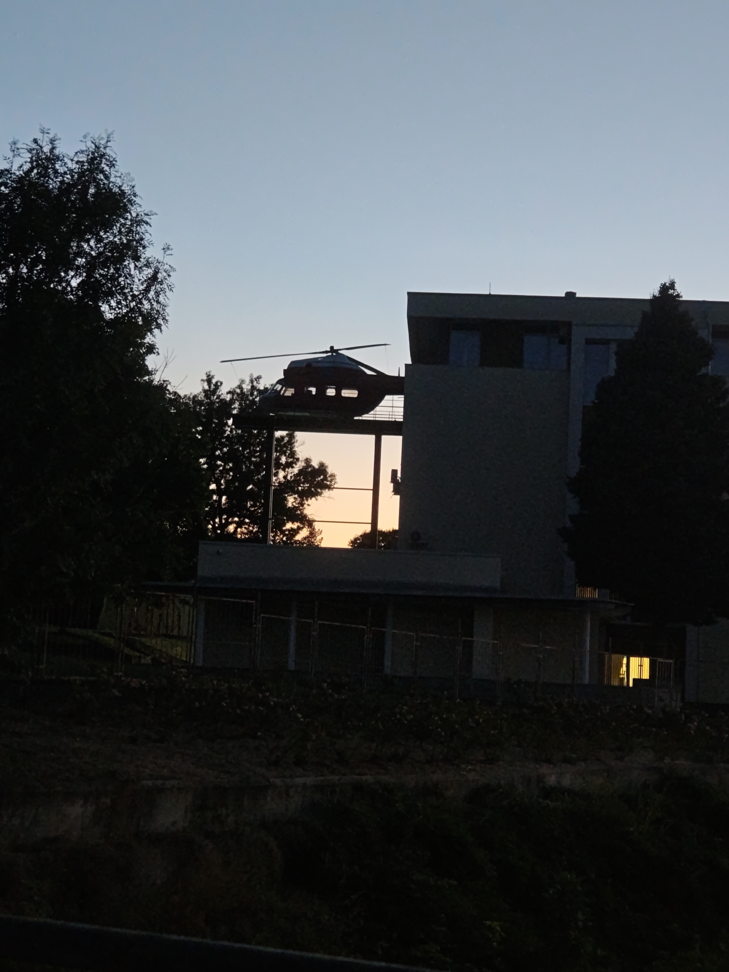 A dark, silhouetted helicopter rests on the roof of a modern building at dusk. The building features large windows that reflect the orange and yellow hues of the sunset. Trees, also in silhouette, frame the scene on the left and right. The overall impression is one of stillness and quiet anticipation. The image is taken from a lower vantage point, looking up towards the helicopter and building. The sky above is a clear, pale blue, transitioning into a deeper dusk color. The location is the city of Pleven, Bulgaria.