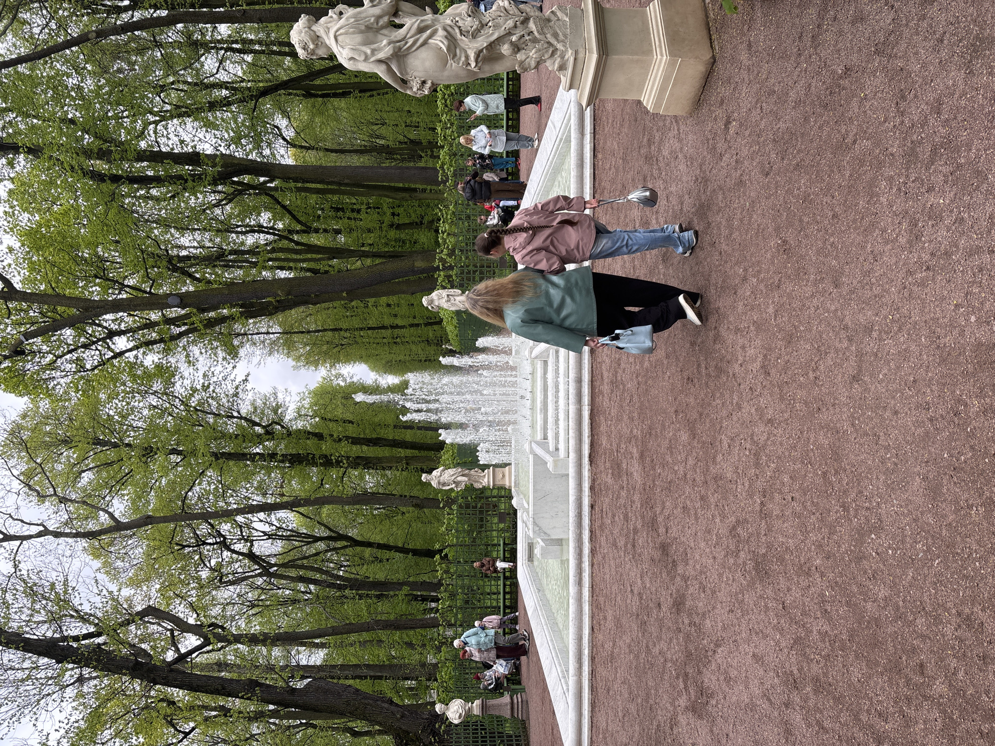 This image captures a scene in what appears to be a park in Saint Petersburg, Russia, likely within one of the historical gardens.
Foreground: A pathway covered in a fine gravel or reddish dirt dominates the lower portion of the image. Two women are walking on the path, towards the right of the image. The woman closest to the viewer is wearing a green jacket and black pants, carrying a light blue purse. The second woman is wearing a pink jacket and blue jeans and she is holding a silver metal object that extends out in front of her.
Middle ground: A long, multi-tiered fountain with white stone borders runs parallel to the pathway. The fountain is actively spraying water. There is a row of statues along the edge of the fountain. A decorative wrought-iron fence separates the fountain area from a grassy area with trees. Several people are visible behind the fence, sitting or walking near the fountain.
Background: The background is filled with tall, green trees. The sky is overcast.
General Impression: The scene suggests a typical day in a historical park or garden in Saint Petersburg, with people enjoying the outdoor spaces and features like fountains and statues.