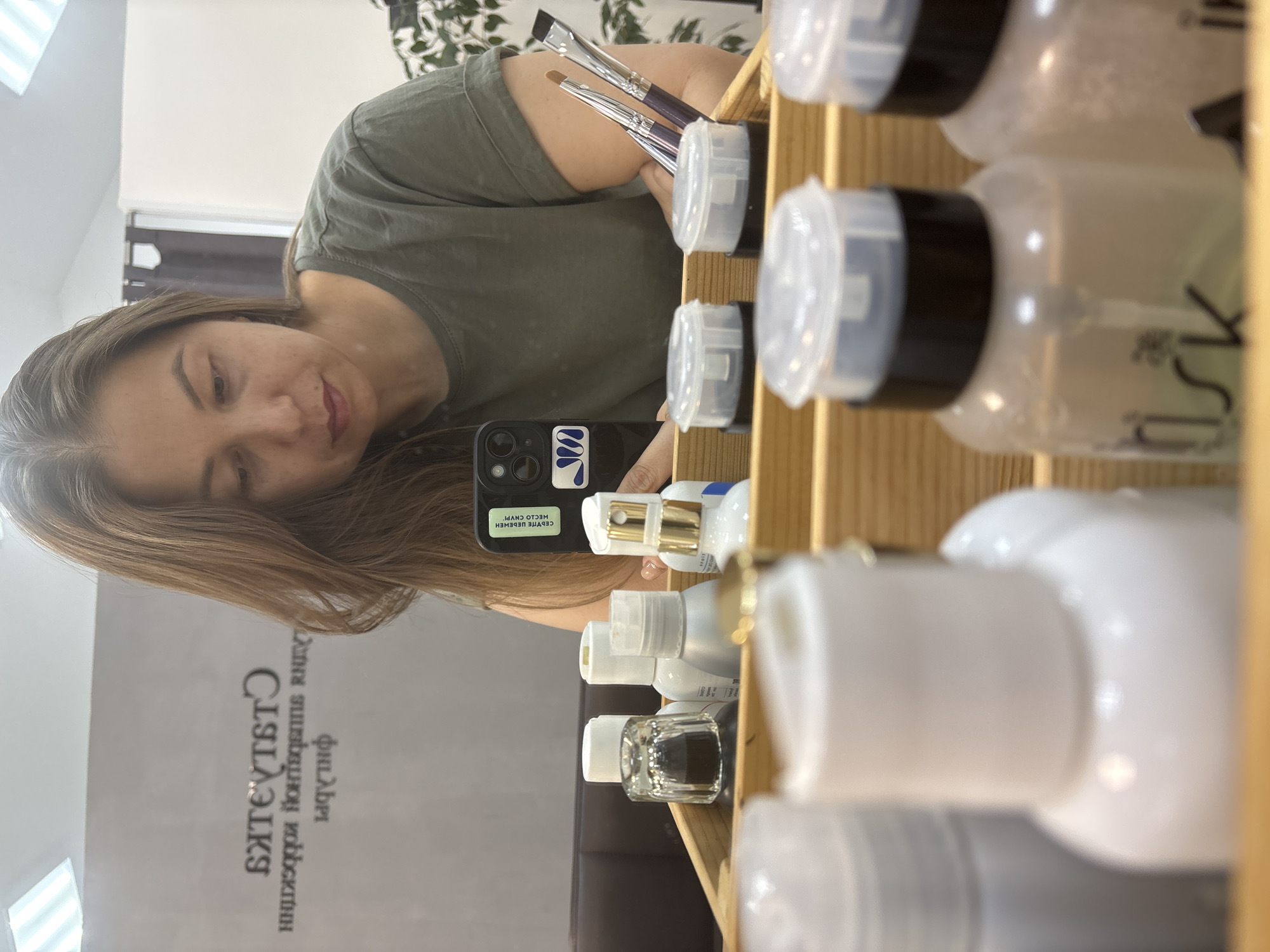 The image captures a mirror selfie of a woman in what appears to be a beauty salon or studio. The woman, with long, light brown hair, wears a green t-shirt and looks downwards with a subtle smile. She holds a black smartphone, likely an iPhone, which has a blue and white abstract sticker and a green sticker on its back. The text on the green sticker, mirrored in the reflection, reads "СДЕЛАЙ ЧЕЛОВЕКА МЕЧТЫ СВОЕЙ!" (Make the person of your dreams your own!). Several makeup brushes are visibly tucked into her right sleeve or held near her arm.
To the right side of the frame, a wooden shelf holds multiple clear and white bottles of varying sizes, containing what seem to be cosmetic or beauty products. Some bottles have black caps, others white, and one features a gold pump dispenser. In the background, a leafy green plant is visible, along with an overhead light fixture. On a dark grey wall, partial Russian text is reflected, including "филибри" and "СТИЛЕТ" (likely part of a salon name related to "Style"). The overall setting, combined with the presence of beauty products and brushes, suggests a professional beauty environment. The lighting indicates it is daytime. The visible Russian text aligns with the location context of Chaikovsky, Russia.