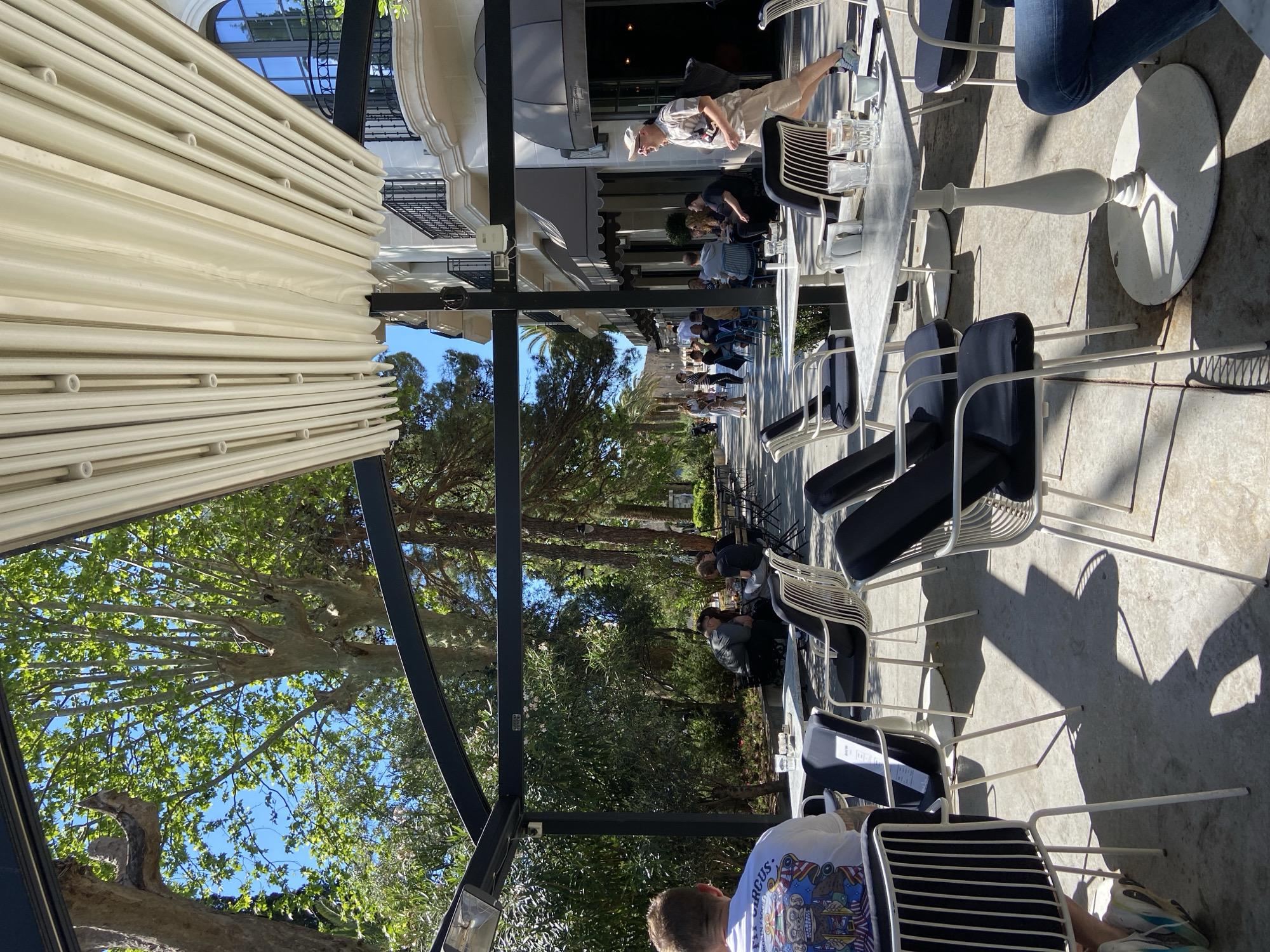 The image captures an outdoor dining area, likely a restaurant or café, in the city of Budva, Montenegro. Several tables with white, marble-like tops and accompanying chairs are arranged on a paved surface. Diners are seated at some of the tables, while others are unoccupied. A retractable awning provides shade over the seating area.
Through the structures of the outdoor seating, a reflected view of buildings and lush trees creates a layered composition. The overall impression is of a relaxed, sunny ambiance in a tourist-friendly location.