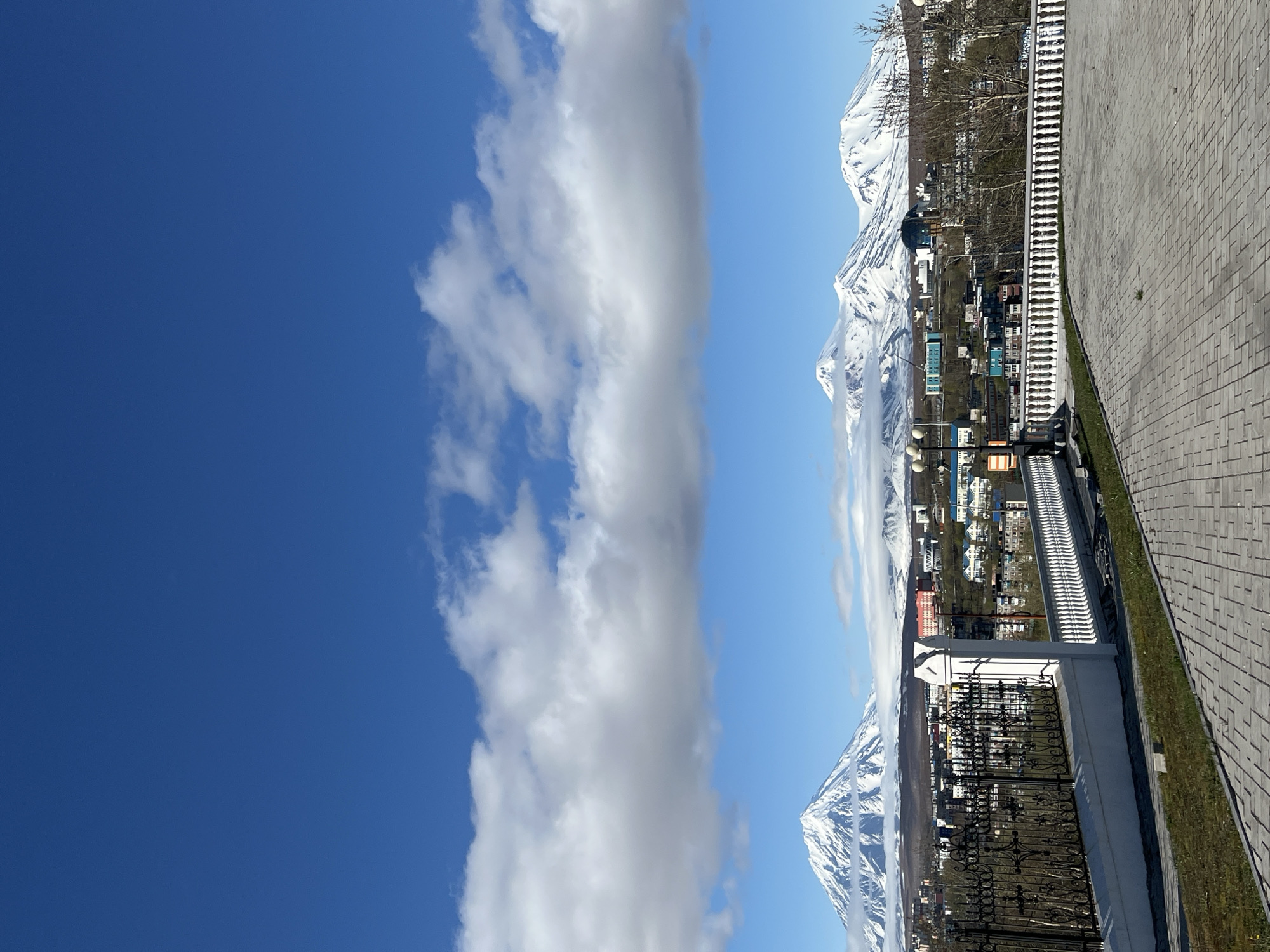The image captures a panoramic view in Petropavlovsk-Kamchatsky, Russia. The sky is bright blue, partially covered by a long vertical stripe of white clouds. Snow-capped mountains, possibly volcanoes, form the background, partially veiled by clouds. A town sits nestled below, with buildings visible amongst sparse trees. In the foreground, there is a wrought-iron fence and a gray brick paved area. The ground is a combination of grass and paved brick.