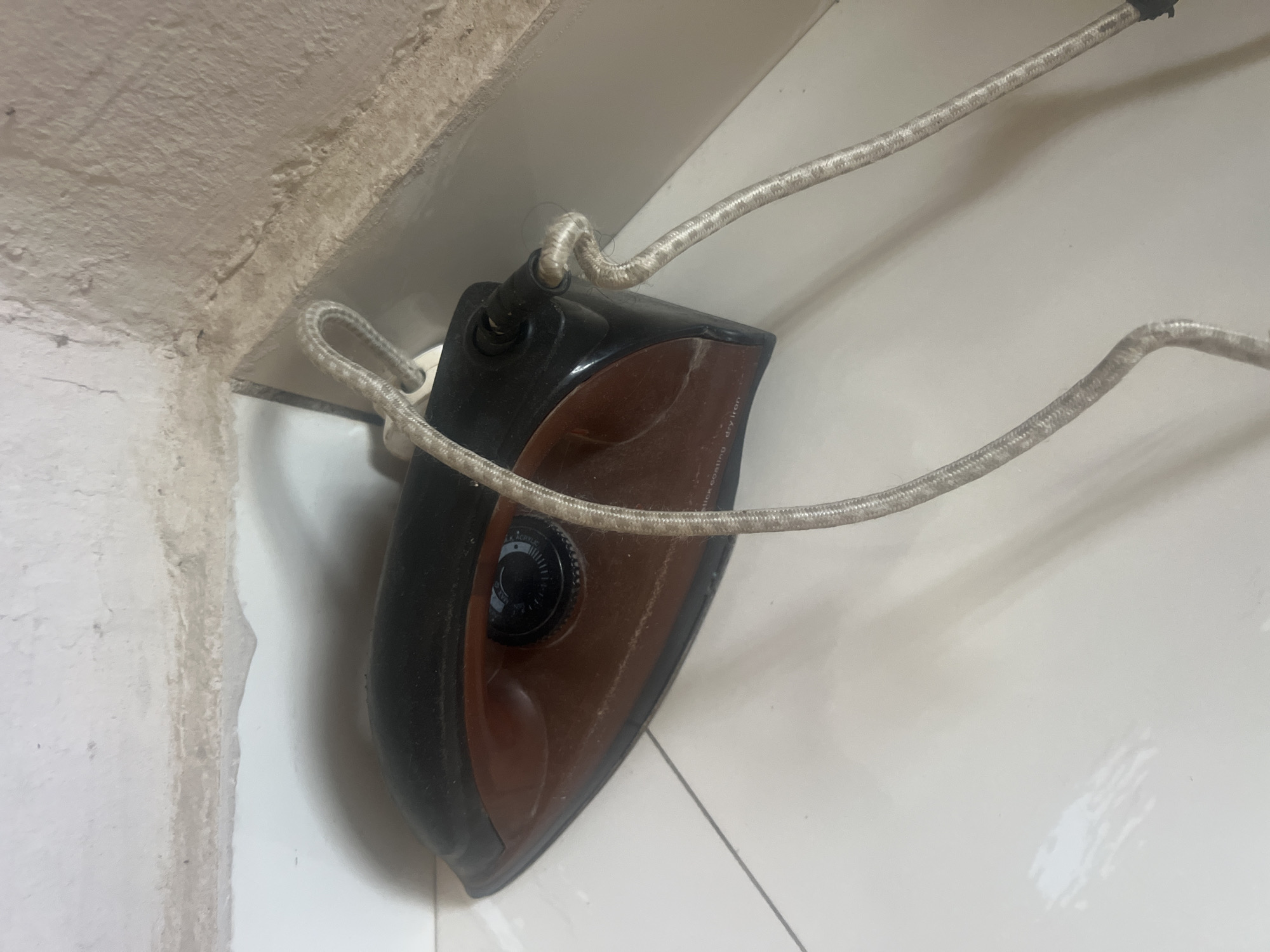 An electric iron is placed on a white tiled floor in the corner of a room, near a wall with rough, light-colored plaster. The iron is black and brown, with a circular temperature control dial on top. Its power cord, covered in a silver woven material, is looped and appears to be tied around the iron's base. The environment suggests an indoor setting, possibly a home in Jalingo, Nigeria, characterized by basic construction materials and a simple tiled floor.