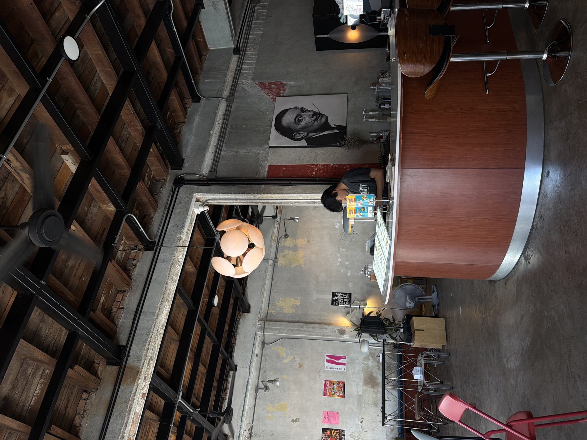An interior shot depicts the counter area of a cafe or bar in Chiang Mai City Municipality, Thailand. The environment features an exposed ceiling with wooden planks and black metal beams, giving an industrial aesthetic. Exposed concrete walls are adorned with various posters and a portrait of Salvador Dali. A person is visible behind the wooden and stainless steel counter, appearing to be working. Several bar stools are positioned in front of the counter. Various objects are present, including bottles, dispensers, a fan, and a decorative pendant light fixture. The flooring is a dark, polished concrete.
