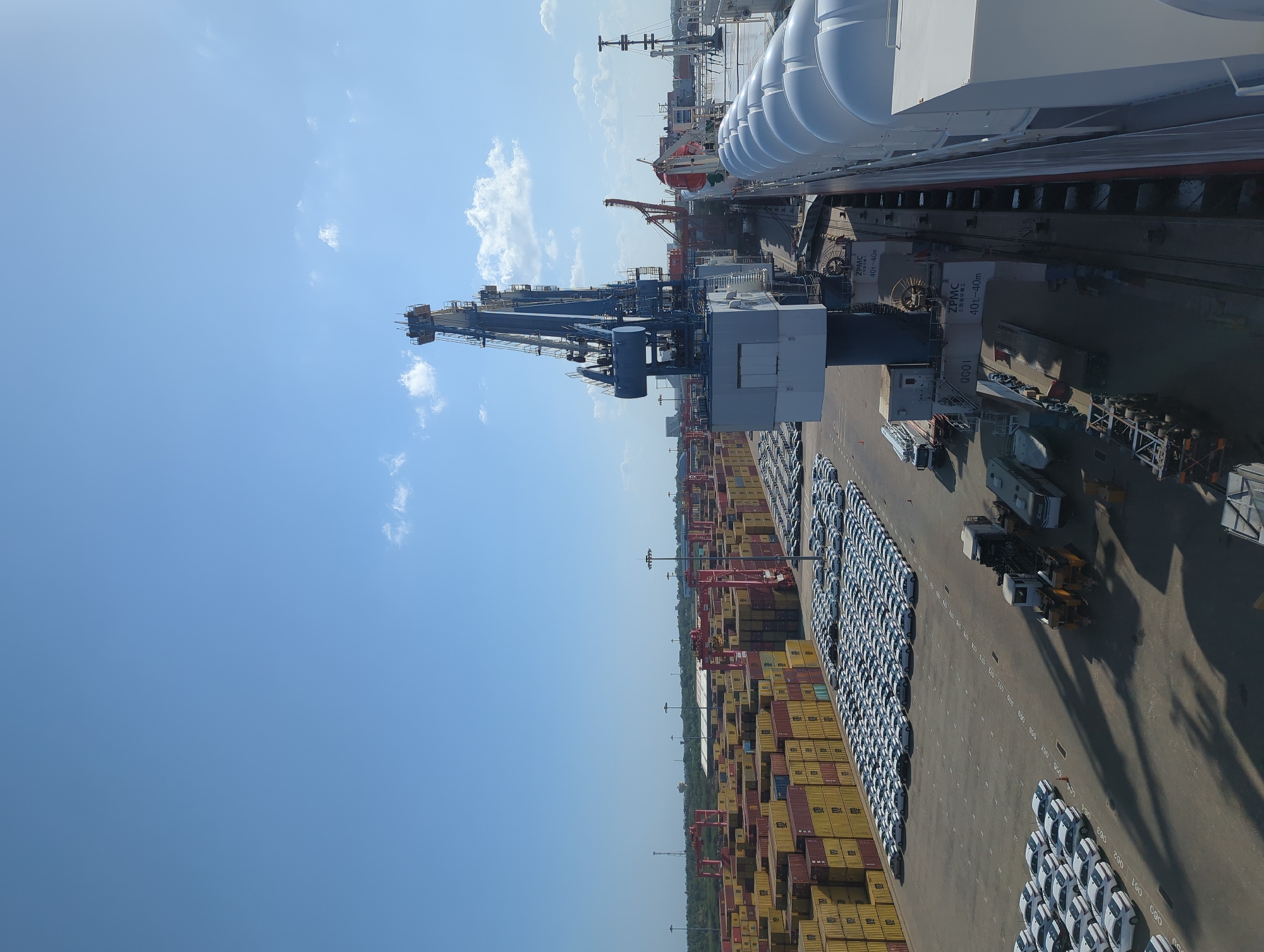 This is a high-angle, vertically oriented photograph taken at the Port of Hambantota in Sri Lanka. The scene depicts a busy port terminal under a clear blue sky with scattered white clouds, suggesting a bright, sunny day.
The primary subjects are large shipping containers stacked high and rows of white vehicles parked on the tarmac. Several industrial cranes are visible, including a large blue gantry crane in the foreground and red cranes further in the background, indicating active cargo handling operations. The presence of the ships, cranes, containers, and vehicles strongly suggests the import or export of goods and automobiles.
Notable details include the text "ZPMC 40t-40m" on some of the crane structures, which likely refers to the manufacturer and capacity of the equipment. The parking area for the vehicles is marked with painted lines and numbers (e.g., "061", "062", "070", "071", "072", "090", "1000"), suggesting an organized system for vehicle storage. The edge of a large white ship, possibly a car carrier, is visible along the top right of the image. The overall impression is one of a functional and large-scale logistics hub.