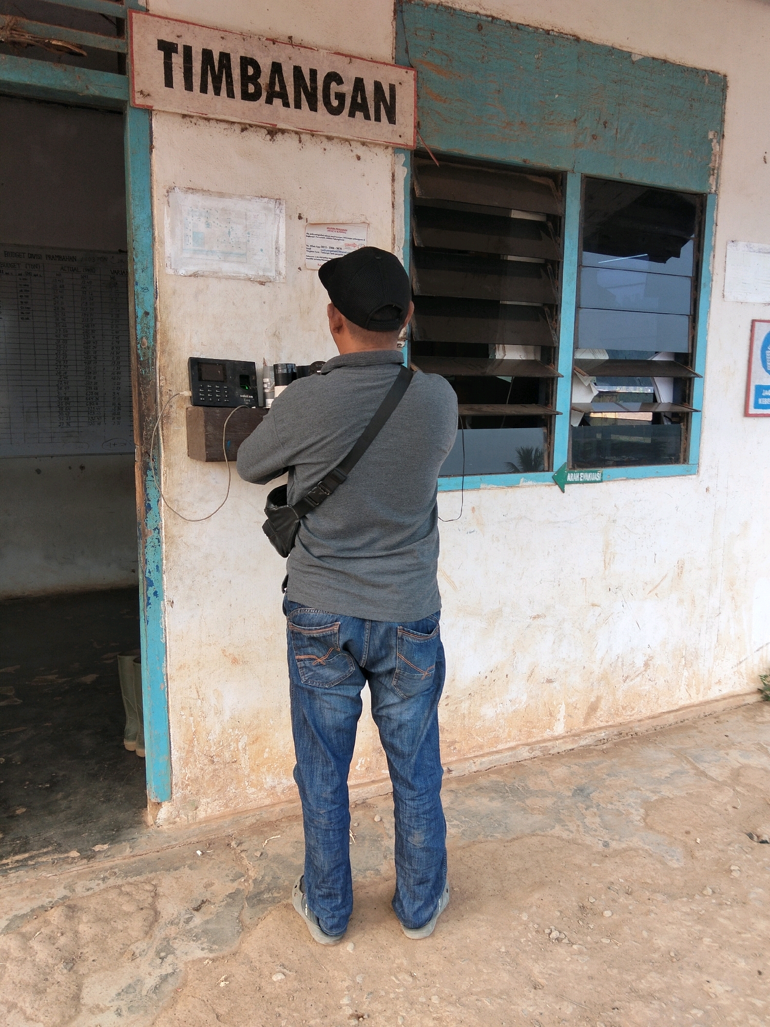 A man in a grey t-shirt and blue jeans stands facing away from the camera, using a black intercom system mounted on a white wall. He wears a black cap and a black bag is slung across his chest. The intercom is connected by a wire to a wooden block on the wall. Above the intercom, a sign reads "TIMBANGAN" in large black letters on a white background. To the right of the sign is a window with blue trim and horizontal wooden blinds, some of which are open, revealing papers inside. To the left of the intercom, a doorway opens to a dark interior with tall green boots visible. The building appears weathered, with peeling paint on the walls and trim. A green arrow sign pointing left with the text "ARAH EVAKUASI" is visible below the window. The ground in front of the building is rough concrete and dirt. This scene is located at the Teluk Tenggirik weighbridge station in Indonesia.