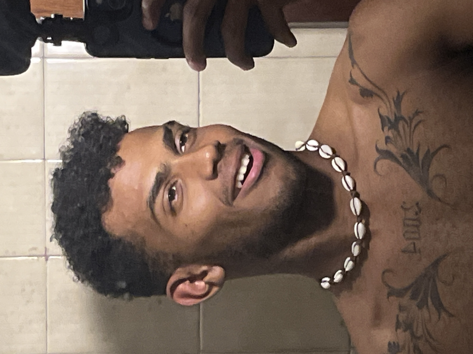 A dark-haired, light-skinned man wearing a seashell necklace is visible from the neck up. He is holding a phone up, seemingly taking a selfie. He has a tattoo above his left clavicle with the year "2007". He is inside a room with tan-colored, square tiles on the wall. No information is provided to determine a location in Porlamar, Venezuela.