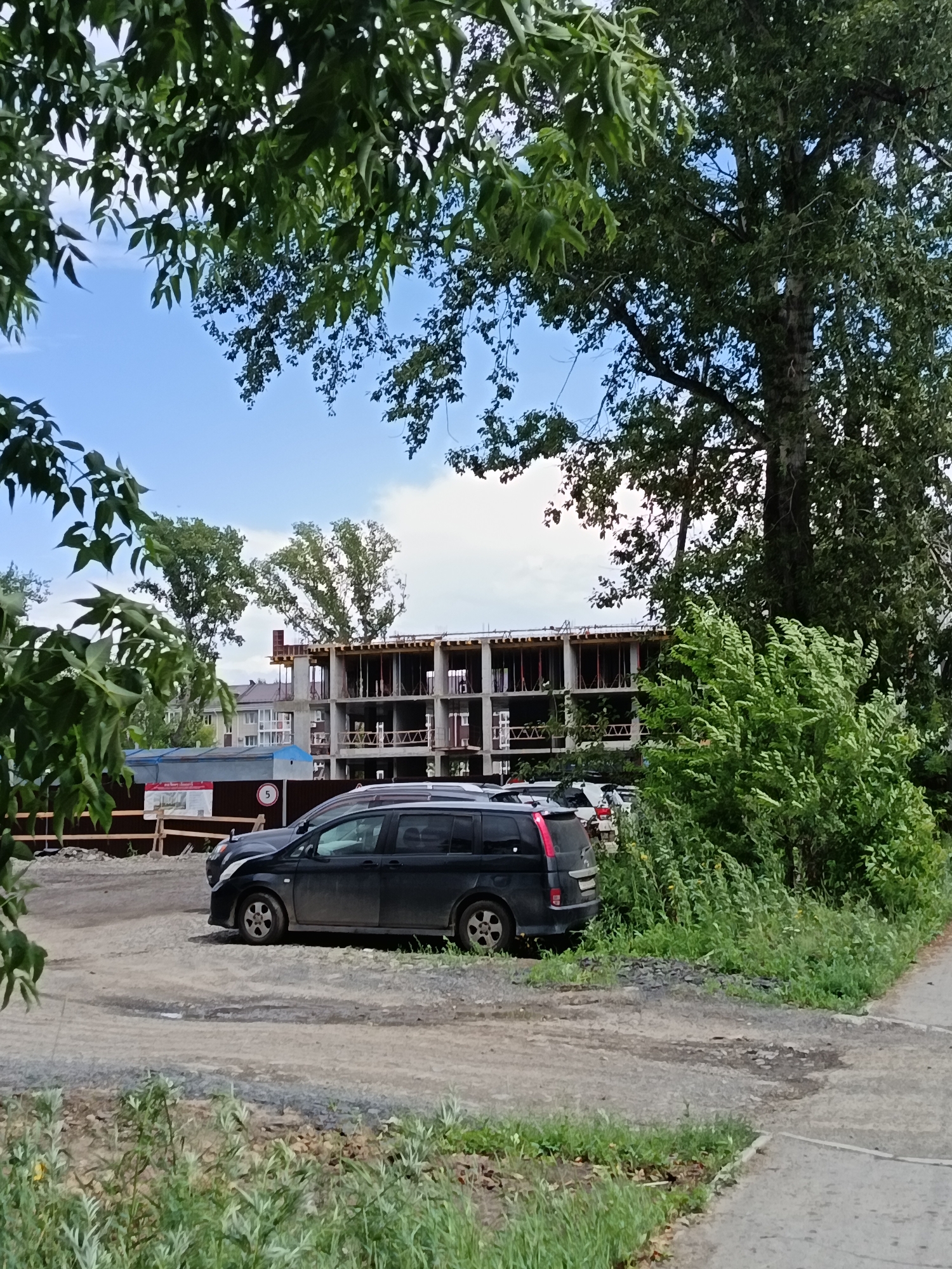 The image captures a construction site in Komsomolsk-on-Amur, Russia, with an unfinished multi-story building at the center. The building is characterized by exposed concrete framework and scaffolding, indicating ongoing construction. In the foreground, a dark-colored minivan is parked on a gravel surface. Surrounding the construction site, lush green trees and bushes dominate the landscape, with some foliage prominently positioned in the foreground, partially obscuring the view. The sky above is a mix of blue and clouds. A wooden fence and a construction sign are also visible. The scene suggests a typical urban development project in progress, set against a backdrop of natural greenery.