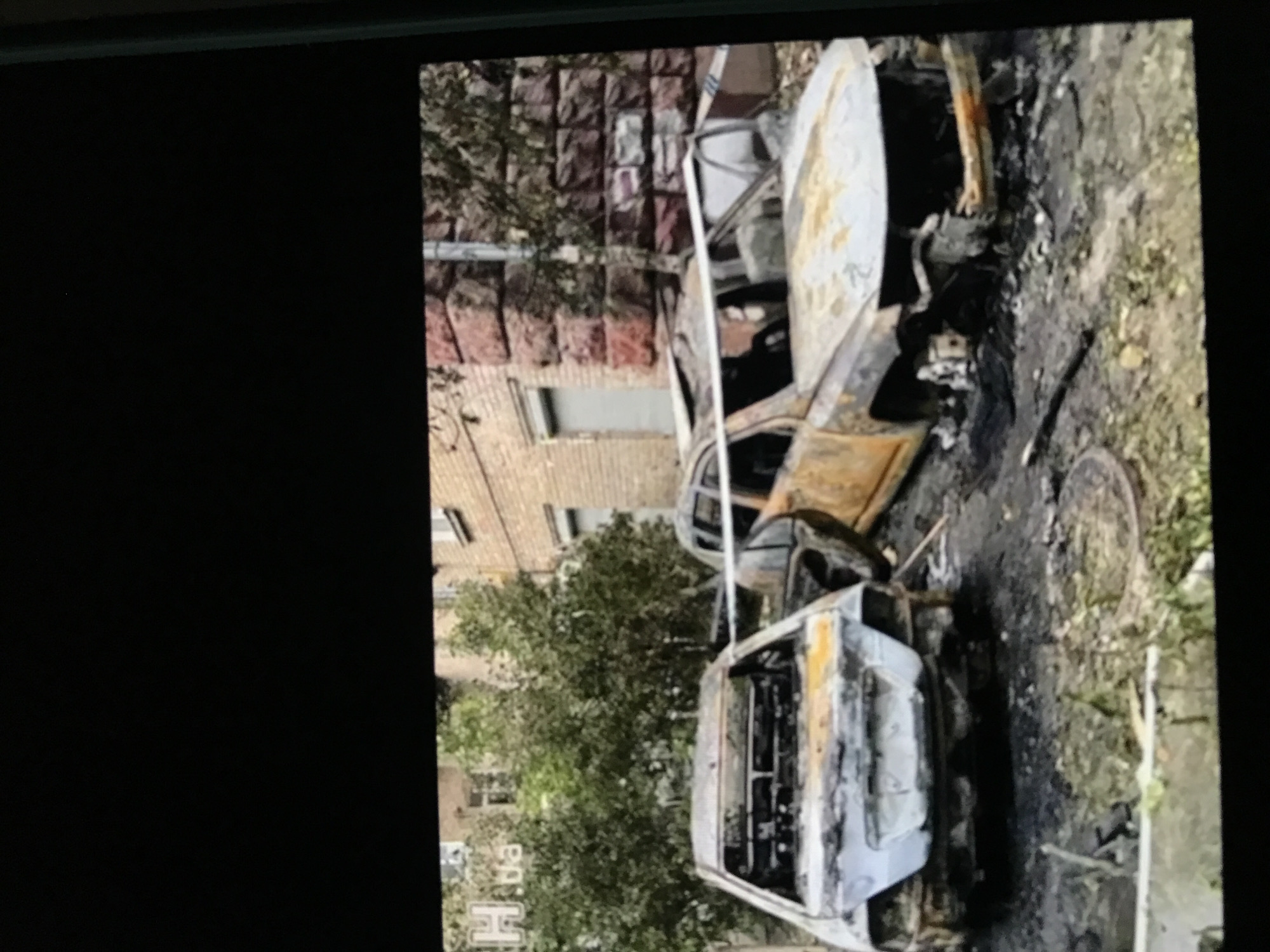 The image depicts two severely burned-out cars on a street in Donji Pobori, Montenegro. The cars are reduced to charred frames, with minimal recognizable features. The street is littered with debris from the vehicles. A building is visible in the background with a brick facade. There's tape strung between the two cars, likely indicating a police or investigative perimeter. A manhole is visible on the ground nearby. The scene suggests a post-fire investigation following a destructive event involving the vehicles.