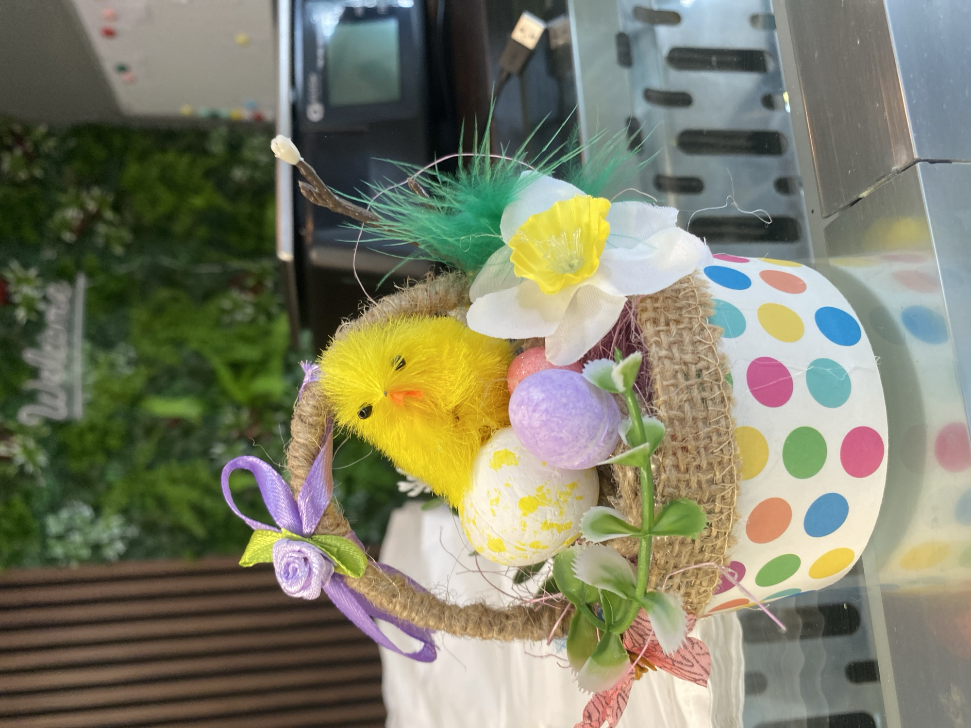 A close-up, eye-level shot showcases an Easter-themed decorative basket. The basket is made of a polka-dotted paper cup, with a burlap band around its rim. Inside, straw is visible, supporting various decorations. A fluffy yellow chick toy is positioned on the left, facing right, with a beak and eyes. Several decorative Easter eggs are placed around the chick: one is white with yellow splatters, another is solid purple, and a partially visible pink egg is also present. Green artificial foliage and a white and yellow faux flower add to the arrangement. A thin twig with a small white bud emerges from the right side of the basket, adorned with green feathers. A purple ribbon with a rose-shaped embellishment is tied to the basket's twine handle. The background is out of focus, showing a wooden-paneled wall on the left and a blurred green, leafy surface behind the basket. A sliver of a black electronic device with a screen and a USB cable is visible on the right side of the background. The scene appears to be set indoors, potentially on a reflective surface, possibly a counter or table. There is no indication of the specific location within the city of Varna, Bulgaria, other than it is an indoor setting.