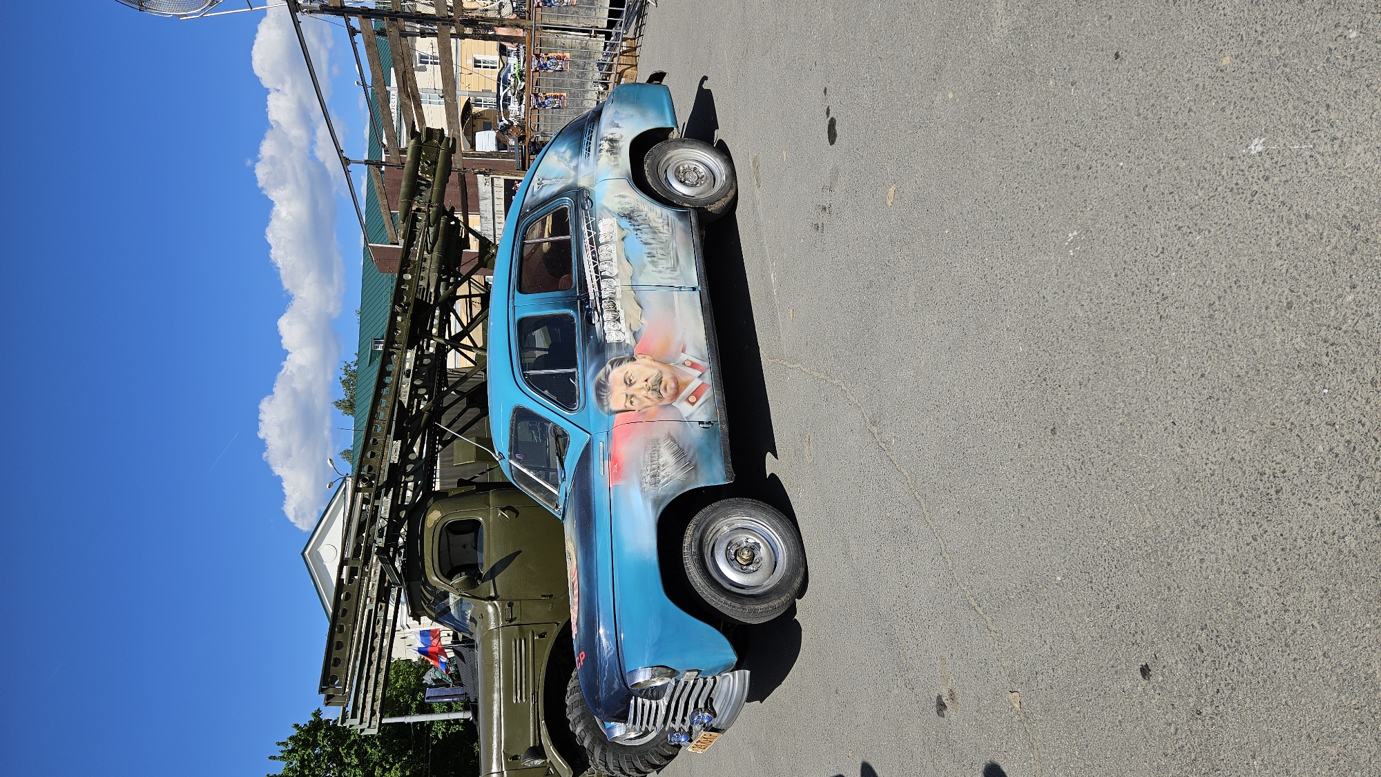 A blue GAZ-21 Volga sedan with a mural of Joseph Stalin and a wartime scene painted on its side is parked on an asphalt surface. The car is positioned in front of a green military truck that has a rack of what appear to be missile launchers on its bed. The truck is parked in front of a building with a green roof. In the background, there are more buildings, some with awnings, and a blue sky with scattered clouds. The scene appears to be outdoors, possibly at a public event or exhibition. The license plate on the GAZ-21 is partially visible and appears to be yellow with black lettering, suggesting it might be a special display plate.