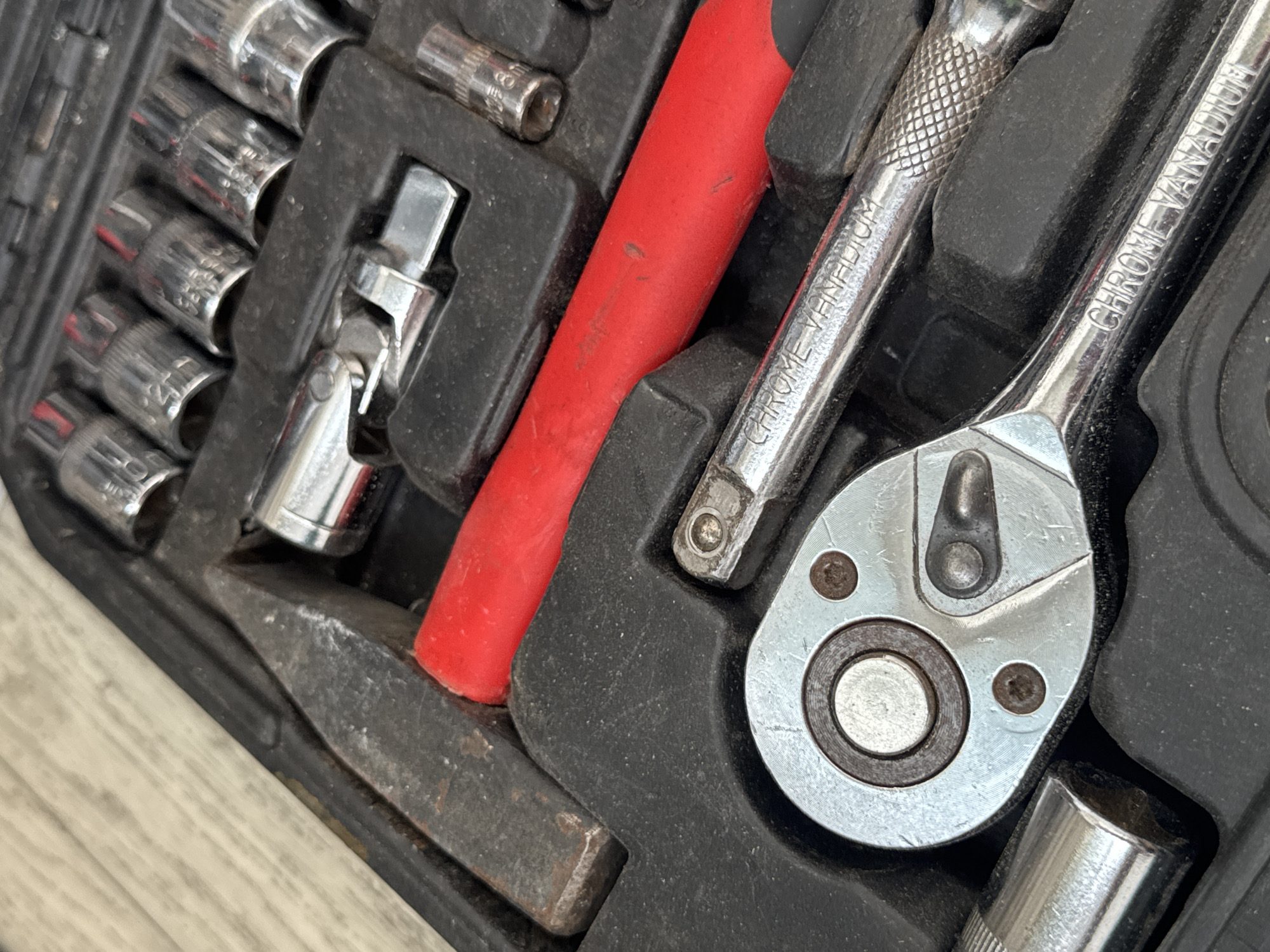 The image displays a close-up, overhead view of a black plastic toolbox containing various metal tools. The tools are arranged in molded compartments within the toolbox. Prominently featured is a silver ratcheting wrench with "CHROME VANADIUM" etched onto its handle. Adjacent to the wrench is a red-handled tool, possibly a hammer, with a dark grey metal head. Several silver sockets of varying sizes are also visible, some stacked and others individually placed in their compartments. The toolbox appears to be resting on a light-colored, textured surface, possibly wood. The lighting is direct, casting some shadows. There are no people or discernible actions taking place in the image. The image is a still life composition focused on the tools within the toolbox. The location is identified as Яромінскі сельскі Савет, Belarus.