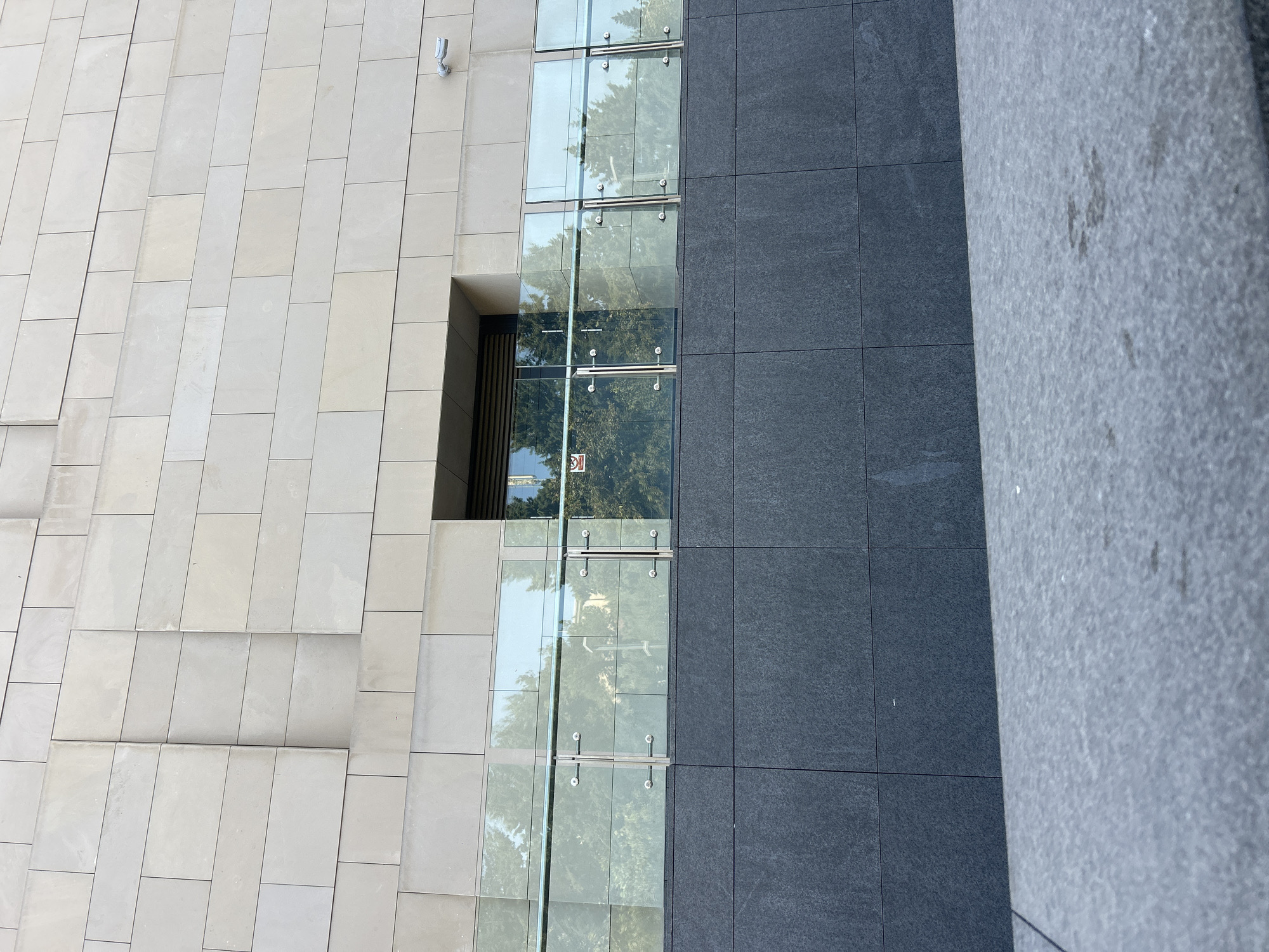 This is a low-angle, full shot of the exterior of a modern building, likely located in Baku, Azerbaijan. The building's facade is composed of large, light-colored rectangular stone blocks in the upper section and dark gray stone blocks in the lower section. A glass railing with metal supports is situated in the middle of the frame, reflecting trees and the sky. Behind the railing, a recessed doorway with a dark frame is visible, and within it, a window with horizontal blinds can be seen. A small, red and white sign is affixed to the glass of the door. A security camera is mounted on the upper portion of the facade to the right of the doorway. The foreground is dominated by a gray, textured surface, possibly a sidewalk or step.