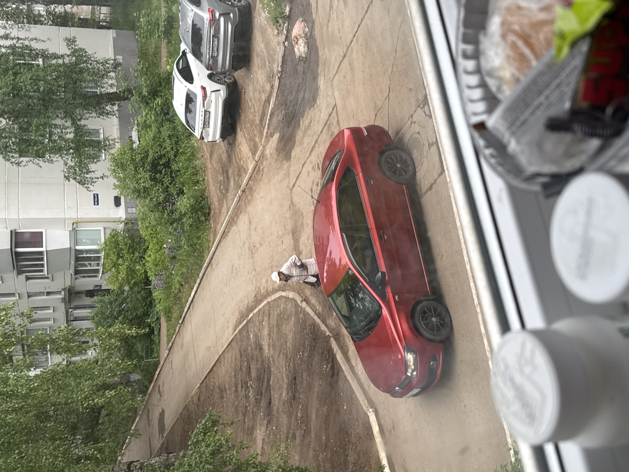 The image shows an outdoor scene from an elevated perspective, possibly from a window. The location is identified as Chaikovsky, Russia, based on the license plates of the vehicles present.
A red sedan is parked diagonally along a concrete walkway, seemingly blocking it. A person wearing a white hat and light-colored clothing is standing near the driver's side of the red car. Other vehicles, a white SUV and a silver SUV, are parked adjacent to each other in the background.
A multi-story residential building with trees in front is visible in the background. The walkway is bordered by vegetation. In the foreground, part of a window frame and objects on a windowsill (containers, wrappers) are visible, indicating the image was taken from inside a building.