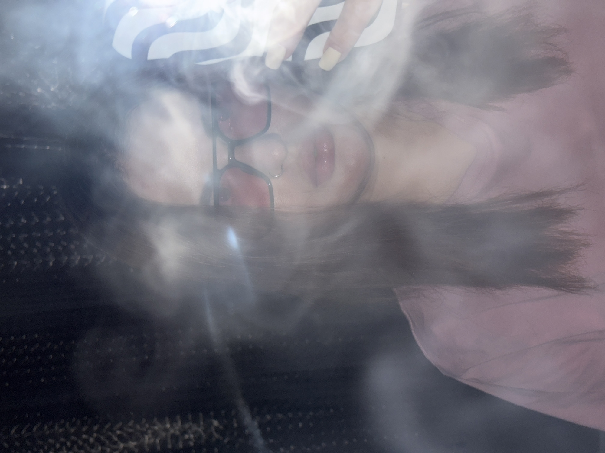 A woman is seen through a blurred and obscured lens, possibly due to smoke or an effect, against a dark background. She wears red-tinted glasses and a light pink shirt or top. Her fingernails are painted a light yellow color. A black and white patterned item is visible near her head. The image does not contain information that supports determining a location or interaction. The location of Krasnodar, Russia, cannot be identified in this image.