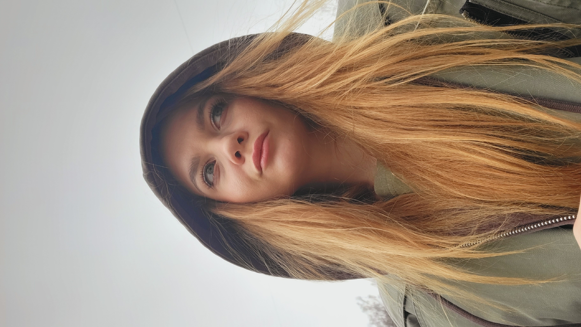 A close-up, low-angle portrait features a young woman outdoors in Chaikovsky, Russia, under an overcast sky. She has long, flowing, light brown or auburn hair that cascades around her face, and light-colored eyes (possibly green or hazel) that gaze slightly upwards and to her left. Her expression appears thoughtful or contemplative.
She is wearing a dark brown hood that frames her face and an olive green, possibly military-style, jacket. A partial silver zipper is visible on the right side of her jacket. The background is predominantly a pale, light gray sky, indicating diffused lighting typical of an overcast day. Faint, blurry outlines of bare tree branches or distant foliage can be seen in the lower part of the background, suggesting a natural outdoor setting. The woman's warm clothing, including the hooded jacket, implies cool or cold weather conditions. No visible text is present in the image.
