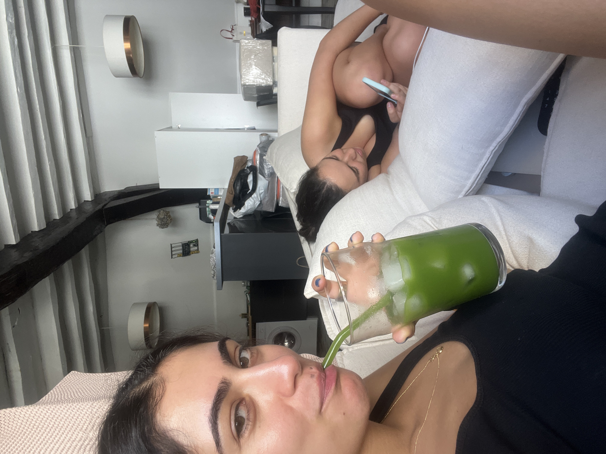 The image shows two young women on a sofa. The woman in the foreground is looking at the camera and drinking a green beverage from a glass. She is wearing a black top and a gold necklace. The second woman is lying on the sofa behind her, looking at her phone. She is wearing a black tank top. The setting appears to be a living room with a white sofa and white walls. There are some decorative items on the wall, including a lamp and what looks like a framed picture. The lighting suggests it is daytime.
