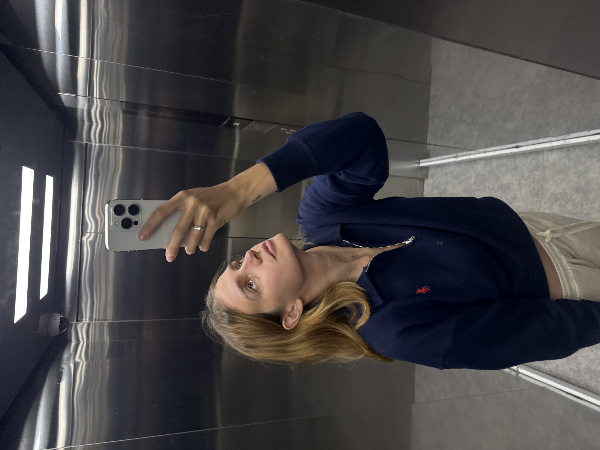 A fair-skinned female is in an elevator taking a selfie. The interior of the elevator is clad in brushed silver metal. The woman is wearing a navy-blue zip-up sweatshirt with a red logo on the left breast. She is wearing a ring on her left ring finger. Her blonde hair is loose and shoulder-length. The floor of the elevator is tiled in a light gray. The location is Unknown, Unknown.