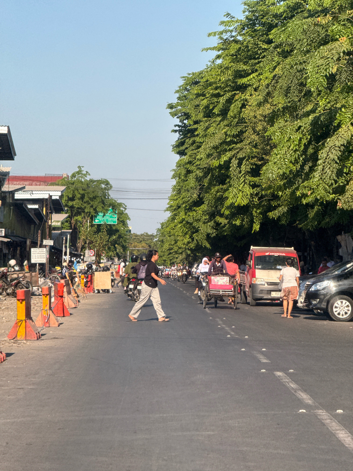The image captures a street scene in Surabaya, Indonesia, likely on a bright, sunny day. The foreground shows a paved road with traffic markings, including white lines and reflective markers. A pedestrian in a black shirt and light-colored pants is crossing the road from right to left, with flip-flops visible on their feet.
To the left of the road, there are construction barriers consisting of orange and yellow traffic cones and poles. Behind the barriers, a row of buildings with visible roofs and awnings lines the street. Several motorcycles are parked along the side of the road.
The middle ground features various vehicles and people. A three-wheeled cycle rickshaw, or "becak," with a pink seat is being ridden by at least two individuals. A red Honda delivery truck is stopped behind the becak. To the right of the truck, a black SUV is parked. A woman wearing a white t-shirt and patterned shorts is standing near the SUV, facing away from the camera.
In the background, more traffic, including numerous motorcycles, is visible. A green directional sign with white text, including place names like "PONDOK DAHIR" and "MADIUN," indicates the city's infrastructure. Large, leafy green trees line the right side of the street, creating a canopy over the road and buildings. The sky is a clear, light blue. The overall impression is one of a bustling urban environment with active street life and ongoing construction.