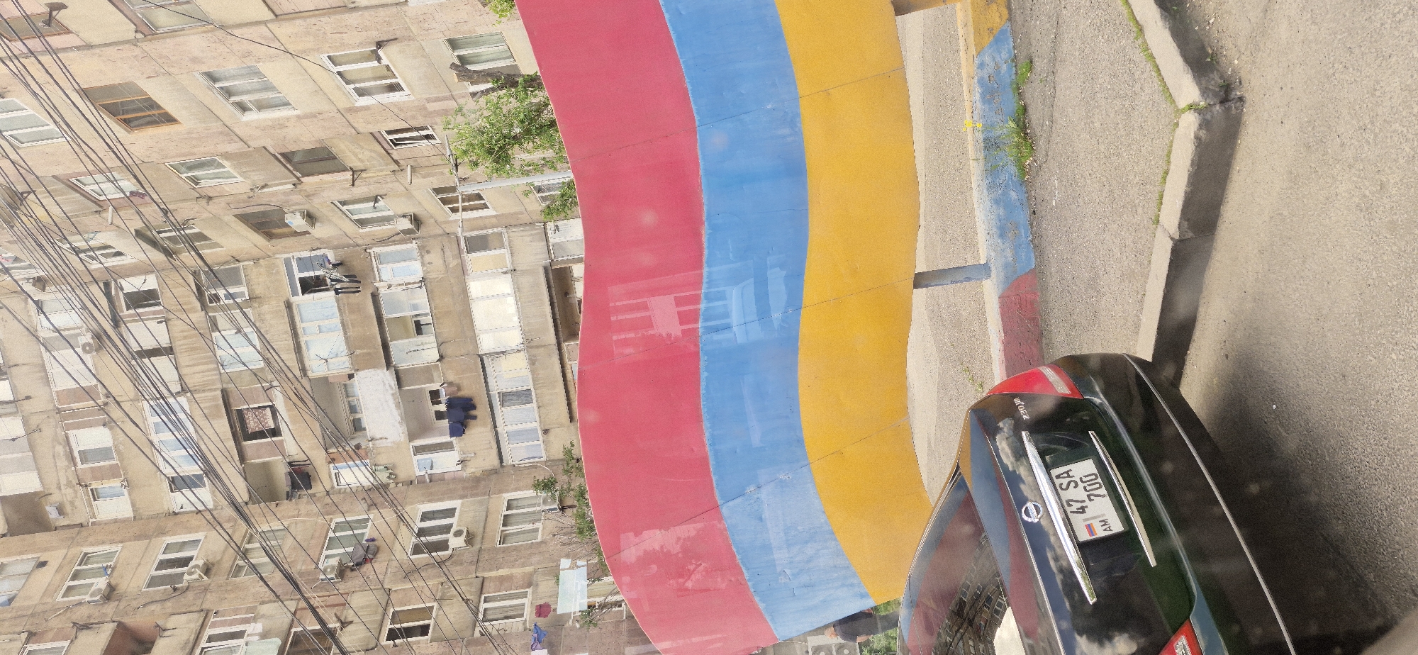 The image shows a building with many windows and power lines in an urban environment. Next to the building is a colorful wall painted in red, blue, and yellow stripes, resembling a flag. A black car is parked near the wall, with the license plate visible, displaying "AM 47 SA 700." The photo is taken from an elevated perspective, looking down at the scene.