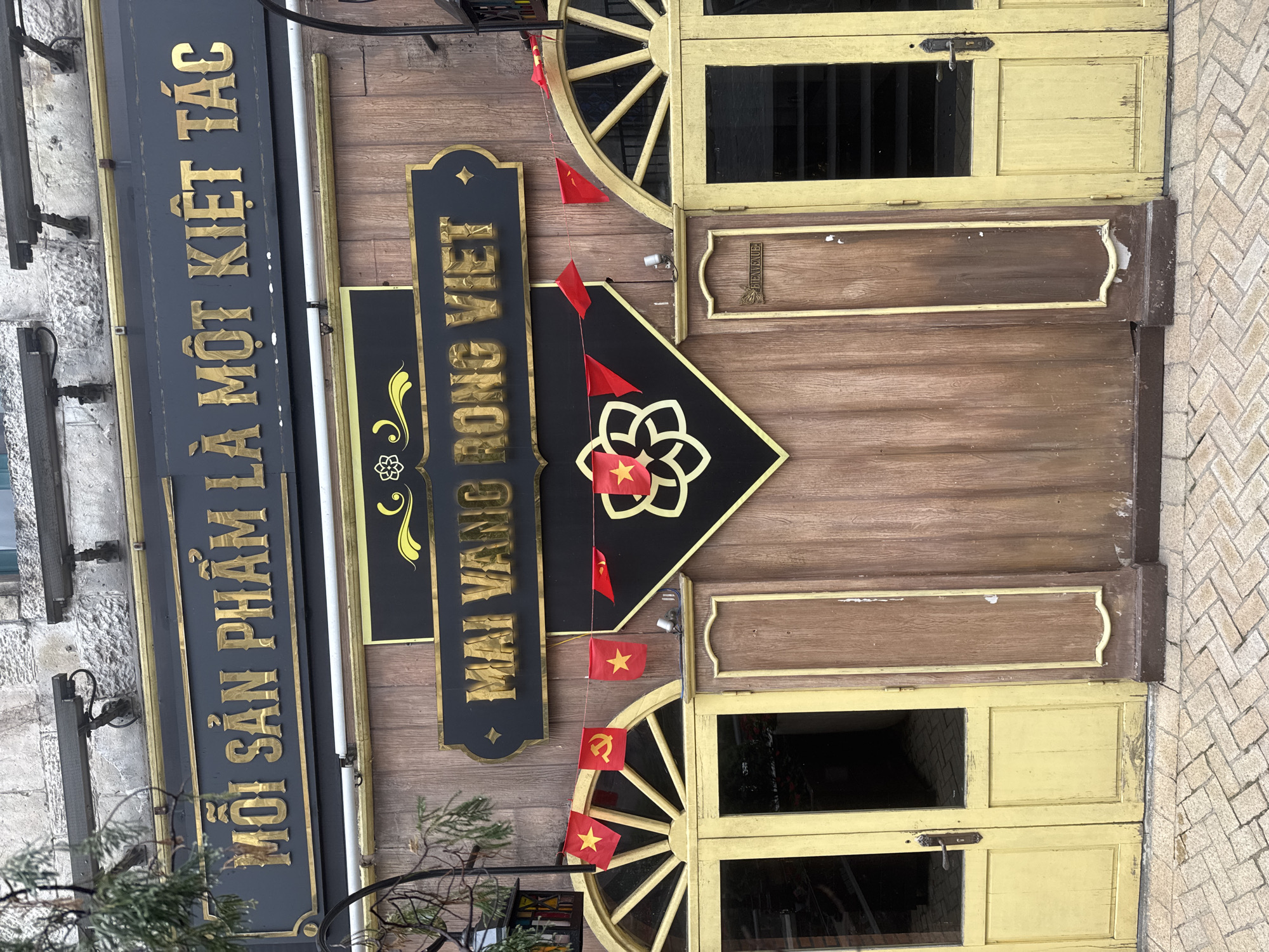 The image shows a building facade, likely a shop, in Đà Nẵng, Vietnam. The building's exterior is predominantly wood with yellow accents. Two distinct wooden doors, adorned with decorative wheel-like designs, are visible. There are Vietnamese signs mounted on the wall, with the text "Mỗi Sản Phẩm Là Một Kiệt Tác" on one sign and "Mai Vàng Rồng Việt" on another. Small Vietnamese flags are draped around the entrance. The building appears to be located on a street with a brick-paved sidewalk.