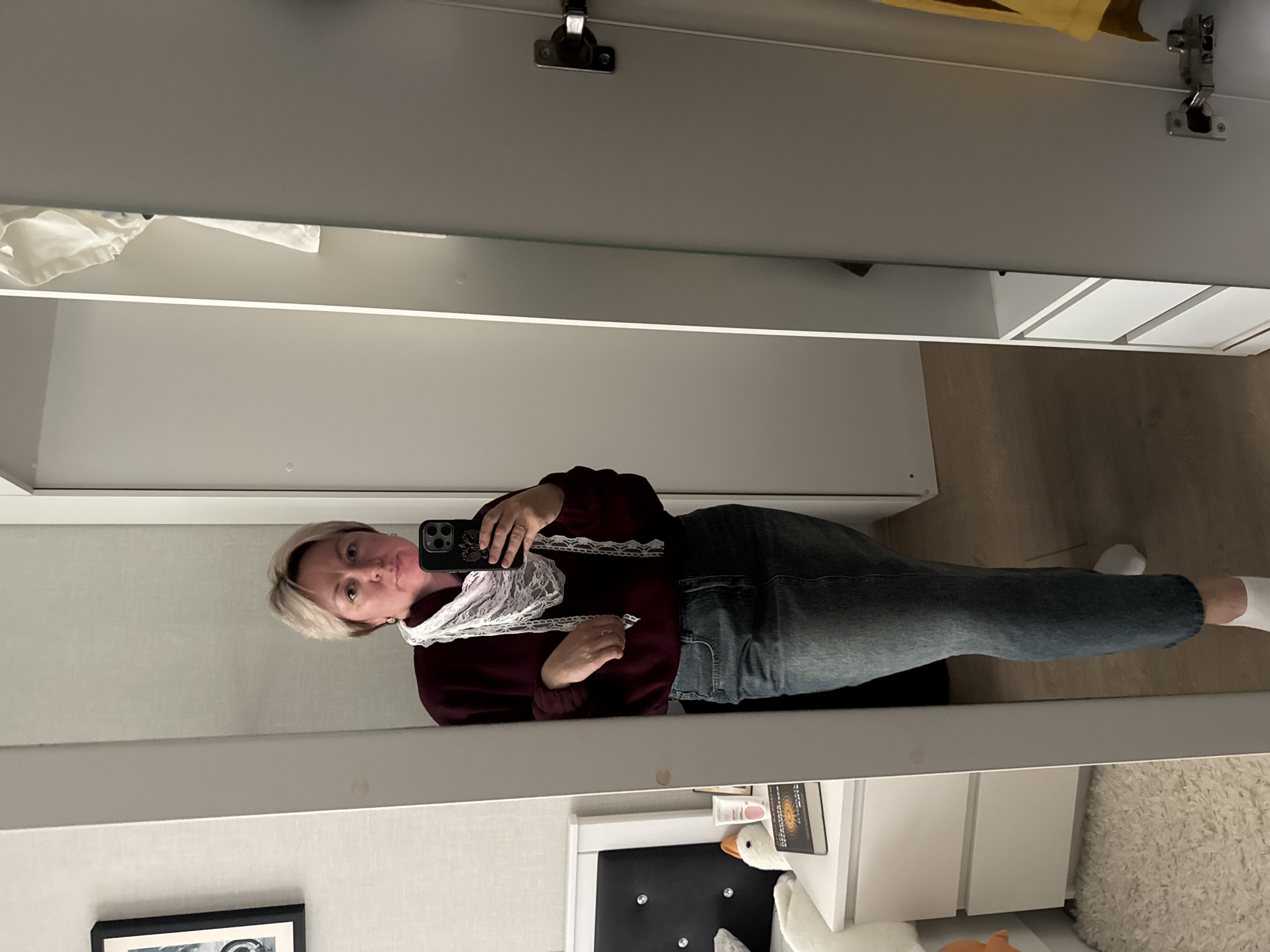 An indoor selfie, rotated 90 degrees clockwise from its original orientation, captures a person reflected in a large mirror. The person, a female with short, light blonde hair, is looking directly at a black smartphone held in her right hand, suggesting she is taking the photograph. She wears a dark red (maroon) long-sleeved top adorned with white lace details around the neckline and chest, paired with blue jeans and white socks. Her expression is neutral with slightly pursed lips.
The background reflected in the mirror reveals an interior setting, likely a room in a home in Chaikovsky, Russia. The walls are light-colored, possibly with a subtle texture. In the upper portion of the mirror's reflection, metal hinges from a cabinet or wardrobe are visible, along with some white fabric. The lower part of the reflection shows a white bedside table or chest of drawers. On its surface, a book with "TAROT" visible on its spine, a white goose plush toy, and a small container that might read "CREME" are arranged. A fluffy white rug lies partially on the floor next to the furniture. A black-framed picture hangs on the wall to the left of the bedside table. The lighting suggests a well-lit indoor environment.