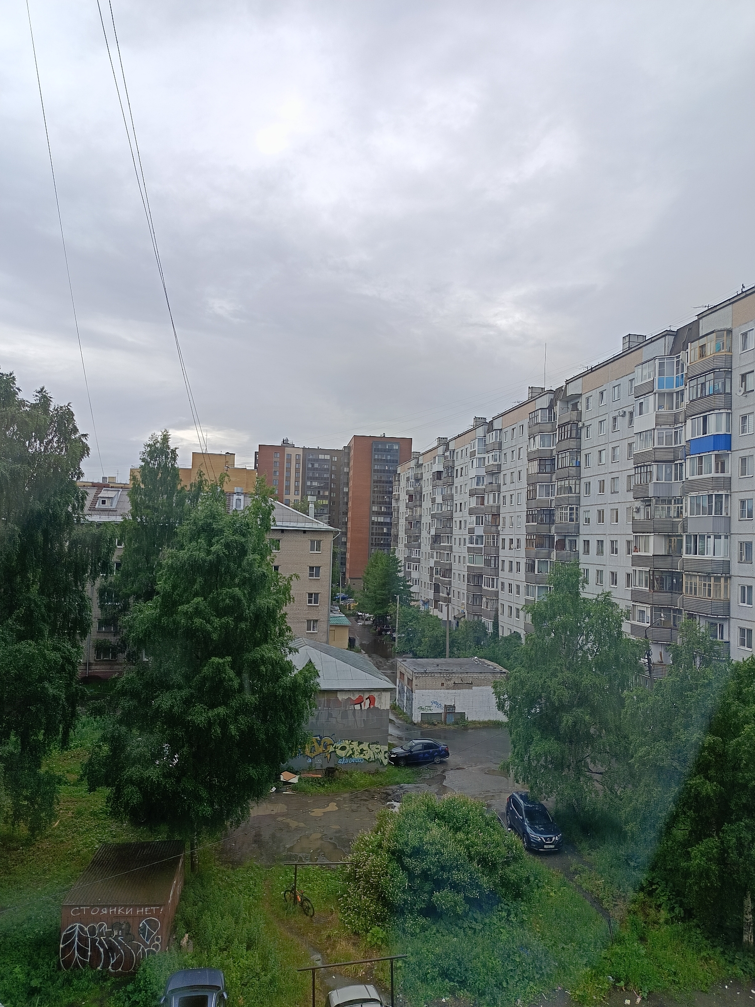 The image depicts an overcast day in a dense urban environment, likely in a post-Soviet country, given the architecture of the apartment buildings. The scene is dominated by a long, multi-story apartment building stretching across the frame, characterized by numerous balconies and a predominantly grey facade. In the foreground, lush green trees and overgrown vegetation suggest a summer or late spring season.
Several cars are visible: a dark SUV parked on a dirt path near the trees, a black sedan further back near a grey building with graffiti, and parts of other vehicles are visible at the bottom of the frame. A bicycle is leaning against a post in the middle ground. A metal garage-like structure in the foreground bears graffiti and the Russian text "СТОЯНКИ НЕТ!", which translates to "NO PARKING!".
The sky is heavily clouded, indicating a gloomy or rainy day, and the light is diffused, suggesting it is daytime, possibly late morning or afternoon. The overall mood is somewhat subdued due to the weather and the utilitarian nature of the buildings. The presence of trees and vegetation softens the otherwise harsh urban landscape.