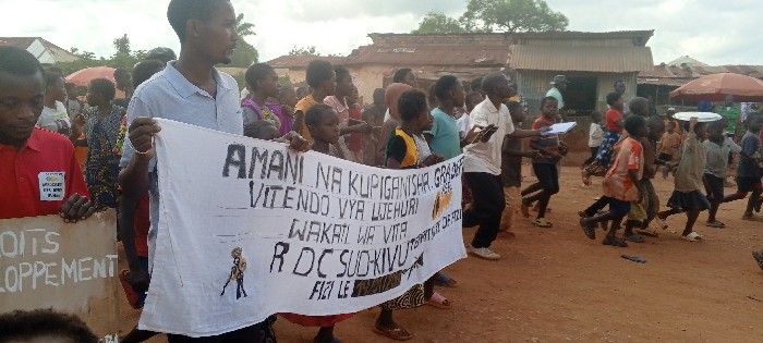 The image depicts an outdoor public gathering on an unpaved, reddish-brown dirt road in the city of Fizi, Democratic Republic of the Congo. Approximately 30-50 individuals, a mix of adults and children, are present, forming a dense crowd that appears to be in motion from left to right.
In the left foreground, a man in a red polo shirt with a yellow badge on his left chest holds a partial brown sign displaying the word "DROITS" and the fragment "LOPPEMENT." Next to him, a man with a beard, wearing a light grey polo shirt and a silver-colored bracelet on his left wrist, holds a horizontal white fabric banner with black handwritten text. The banner reads: "AMANI NA KUPIGANISHA GRAGSE," "VITENDO VYA LWEHURI," "WAKATI WA VITA," "R DC SUD KIVU," "TERRITOIRE DE FIZI," and "FIZI LE." A stylized drawing of a person is positioned below "R DC SUD KIVU." Several children and young people are also visible holding sections of this banner.
To the right of the banner, a man in a white collared shirt holds a white clipboard or book. A group of children are actively moving forward in this area; one child carries a white circular object, possibly a plate or lid, on their head.
The background consists of various single-story structures with corrugated metal roofs, some showing signs of rust. These buildings appear to be of simple construction. Green foliage is visible in the far background. A red parasol is also present in the right background. The sky is overcast.